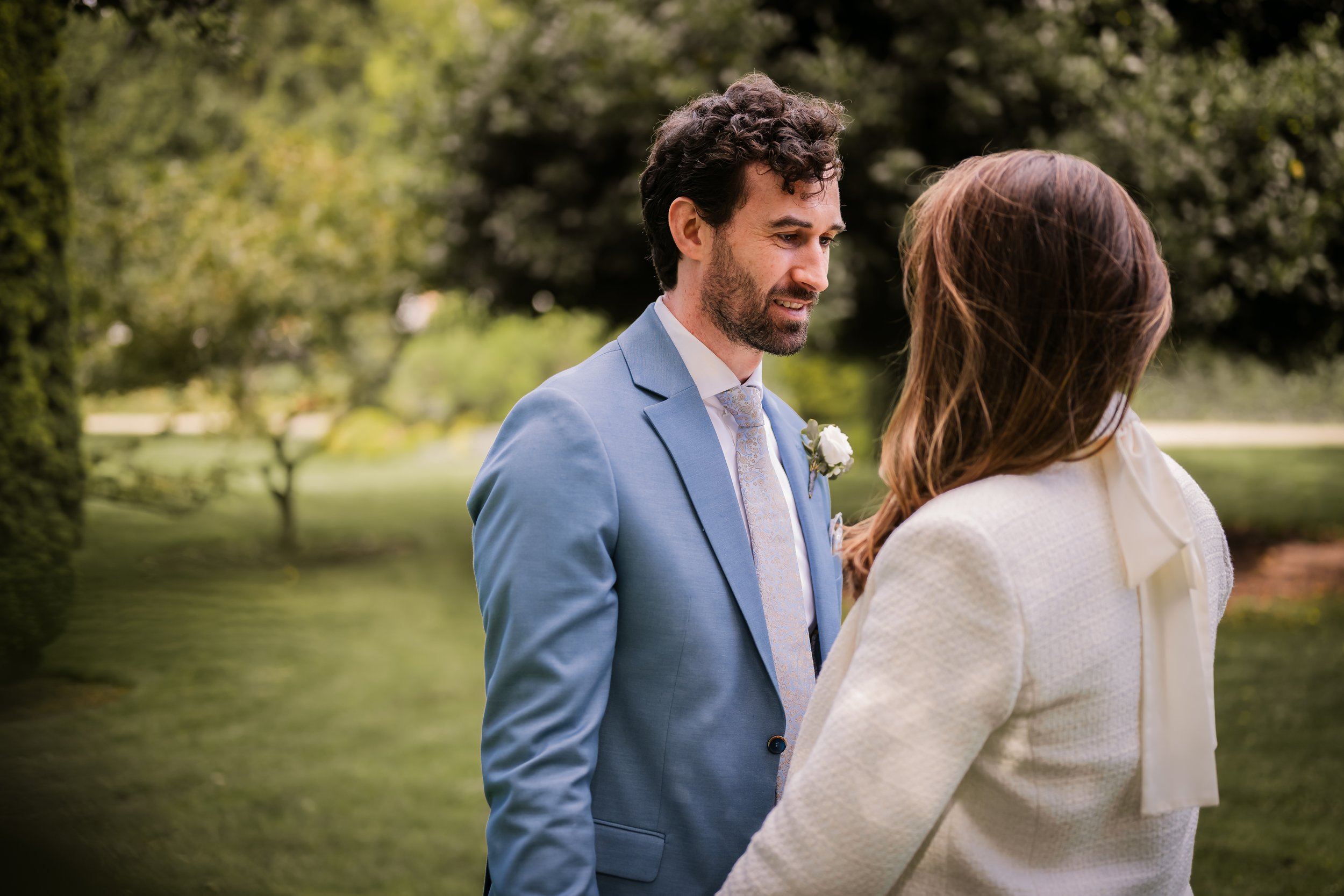 A groom in a light blue suit and a bride in a white dress look at each other outdoors by a pond with trees in the background.