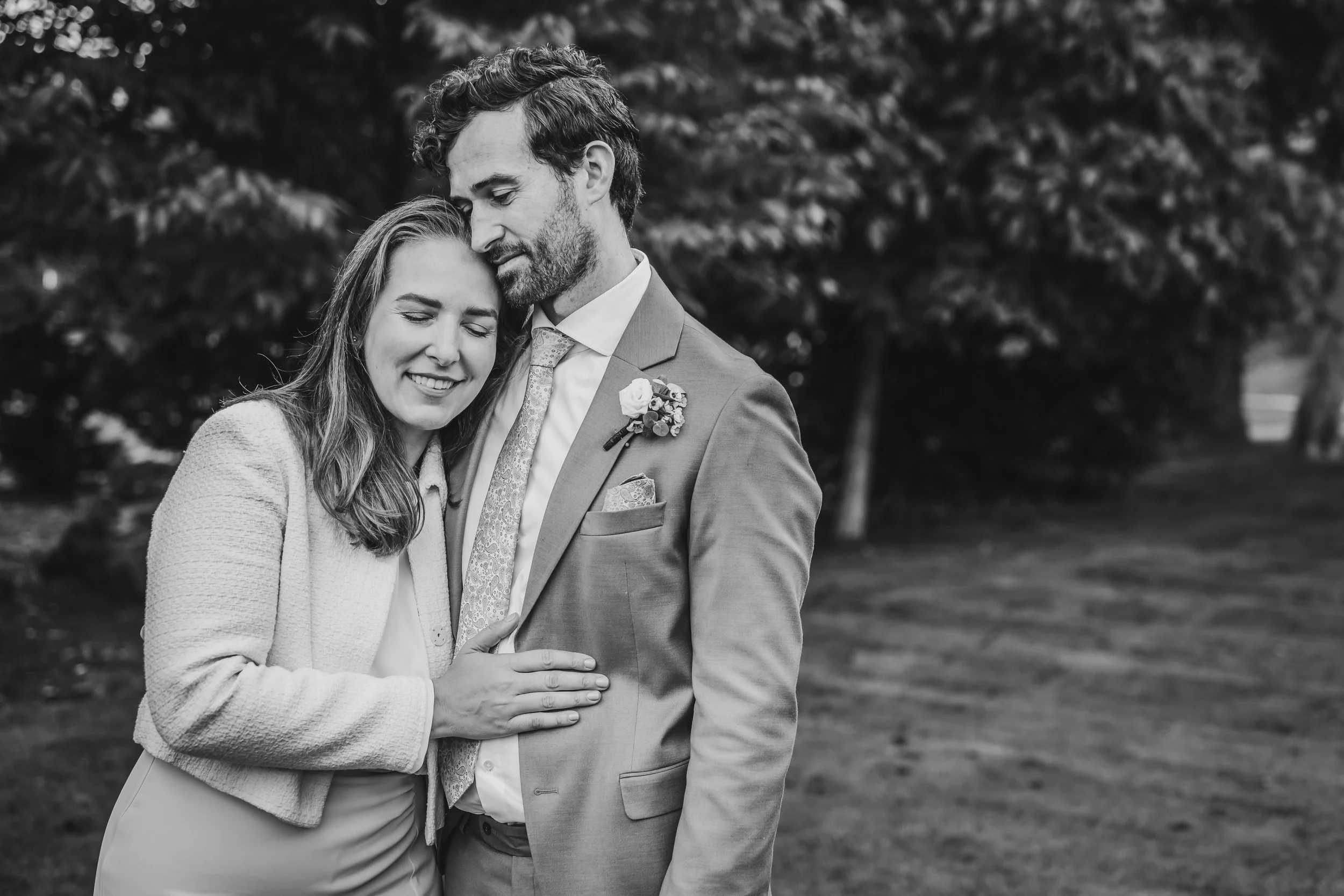 Black and white photo of a happy couple embracing outdoors, with the woman smiling with eyes closed and the man gently holding her, surrounded by trees.