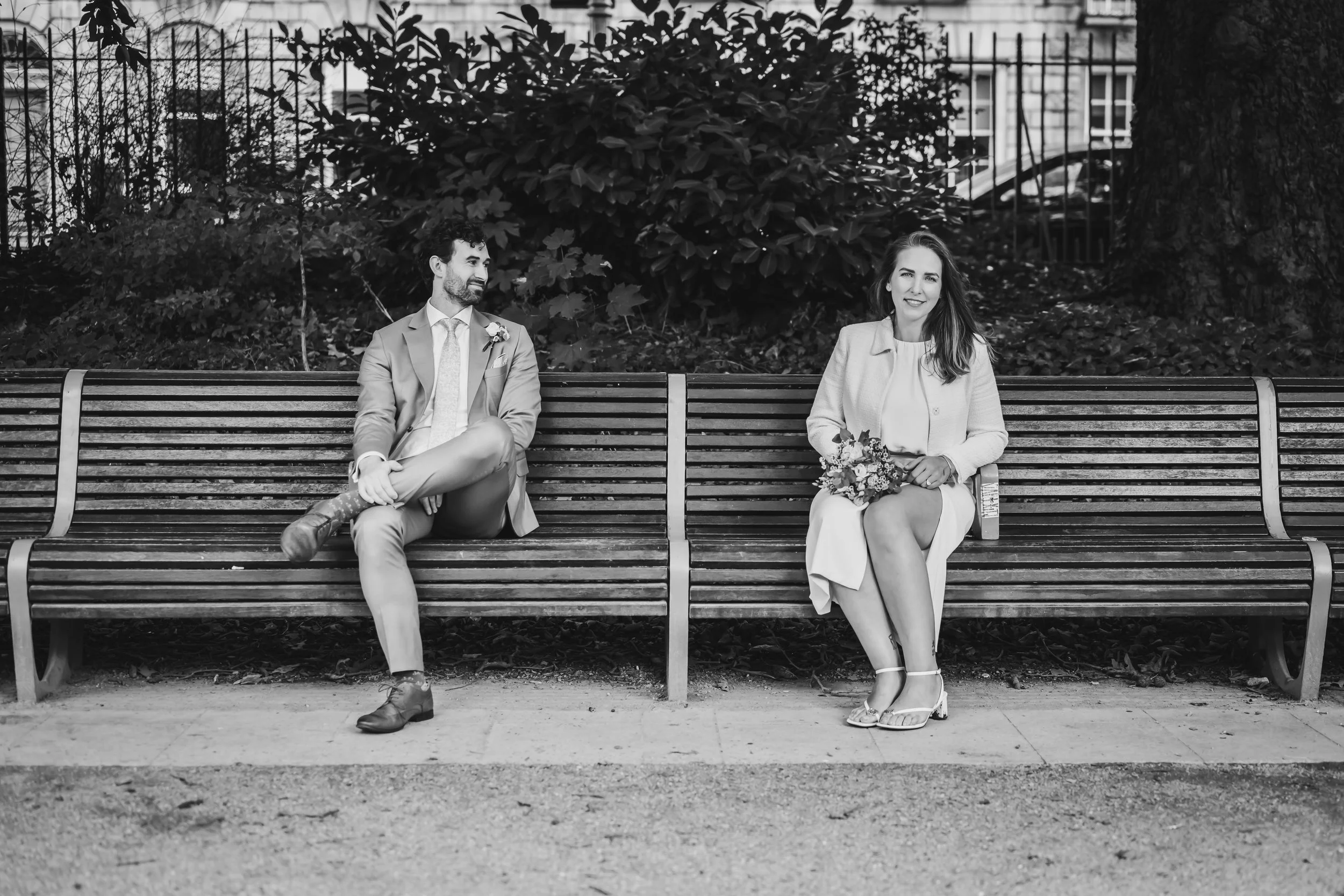 A black and white photo of a groom and his bride sitting on separate benches in a park. The man is looking at the woman, who is holding a bouquet of flowers and smiling.