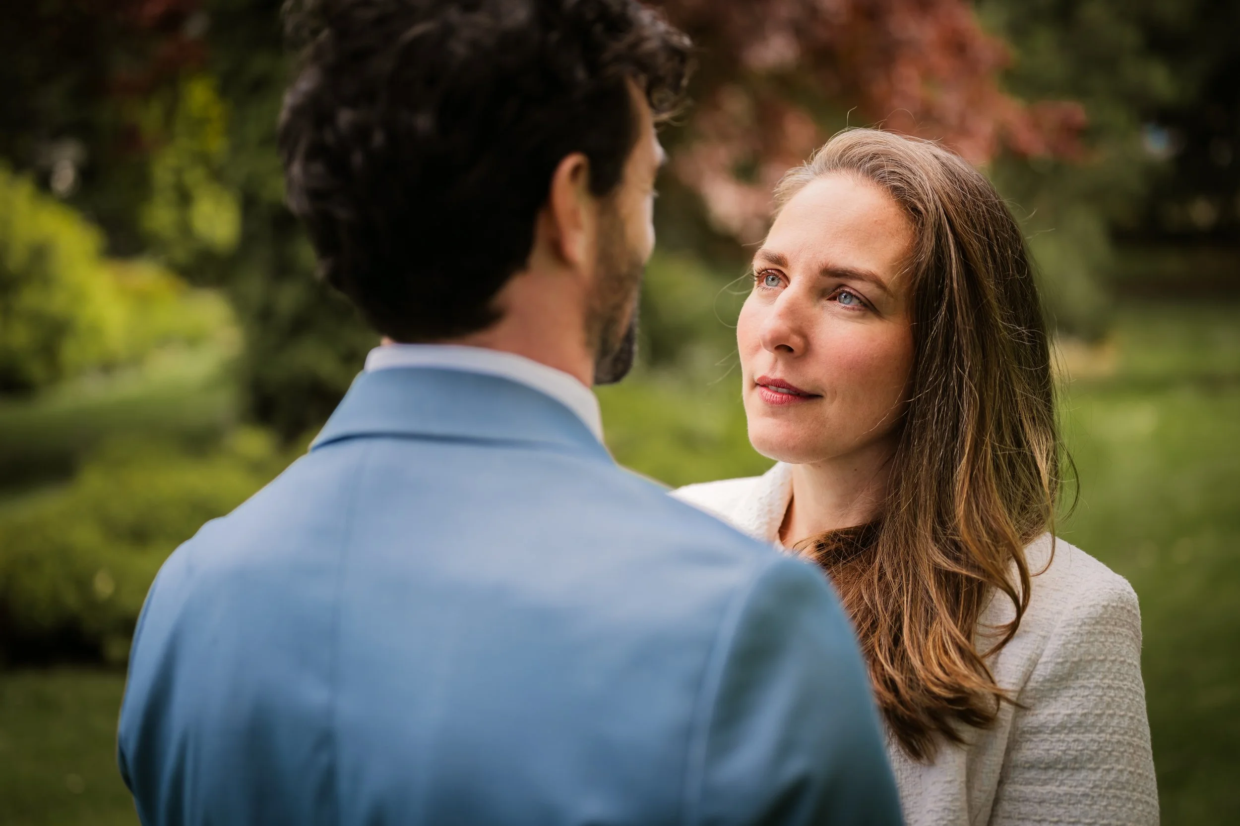 A couple in love face each other outdoors in a lush green park, with the woman looking intently at the man.