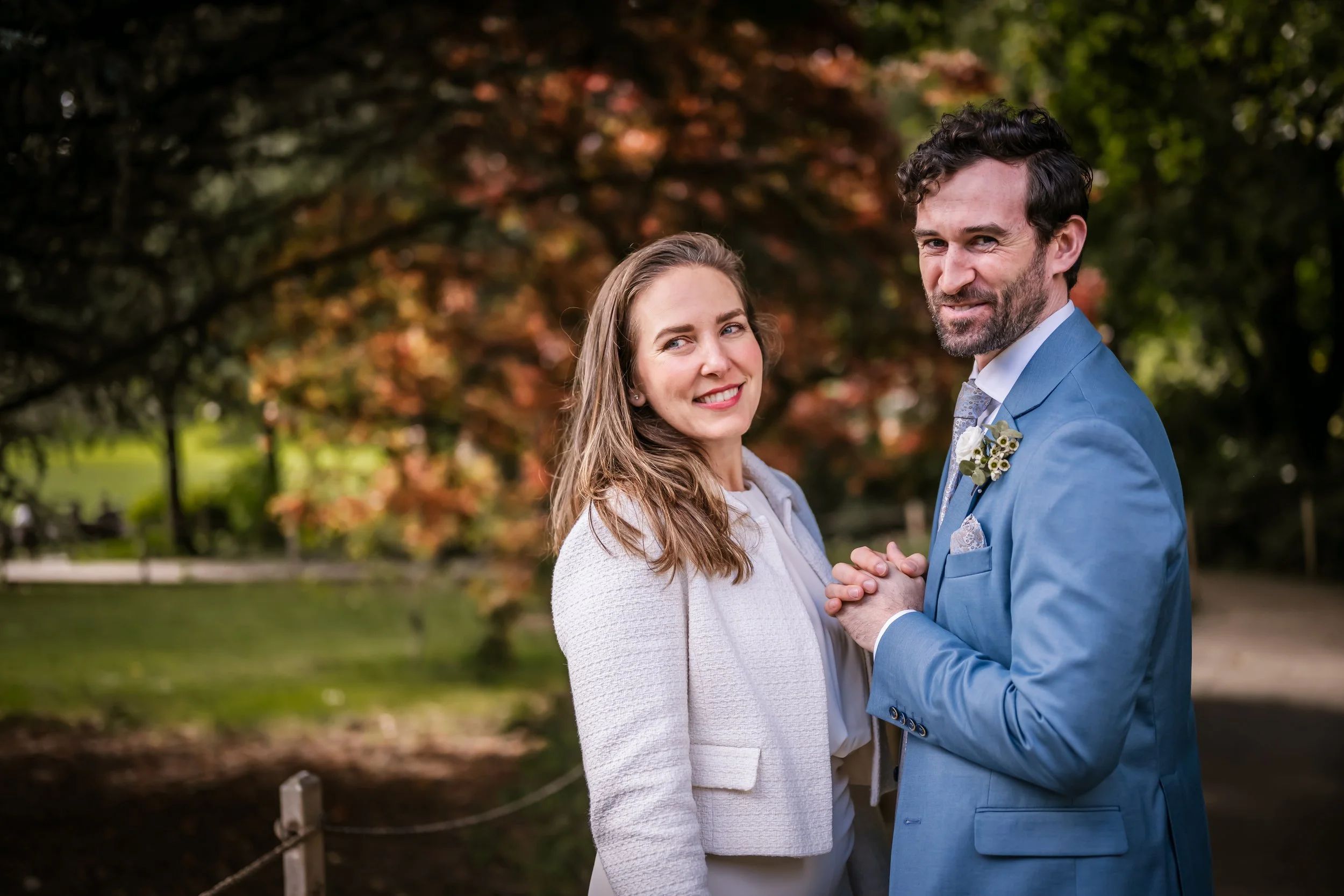 A smiling bride and groom holding hands in a park with autumn foliage in the background.