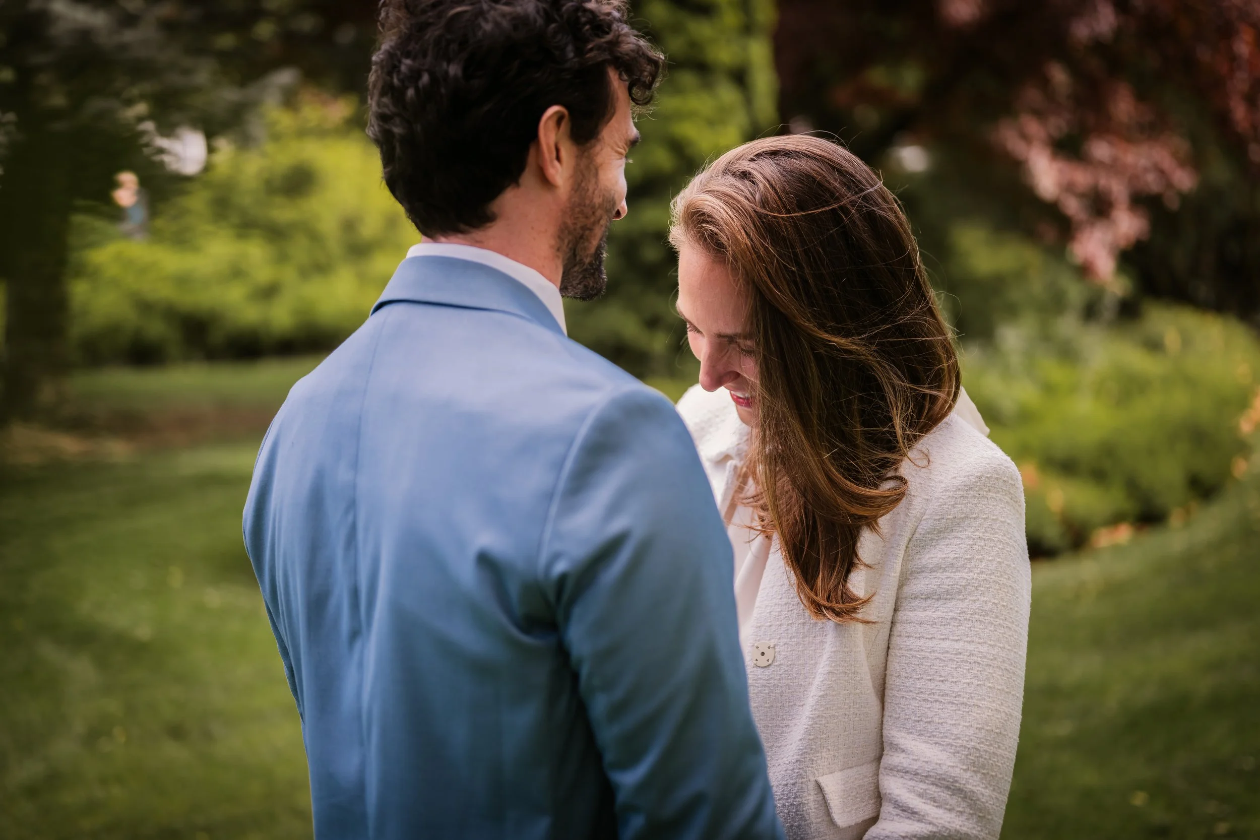 A man and woman share an emotional moment outdoors, with their foreheads touching and eyes closed, surrounded by greenery.