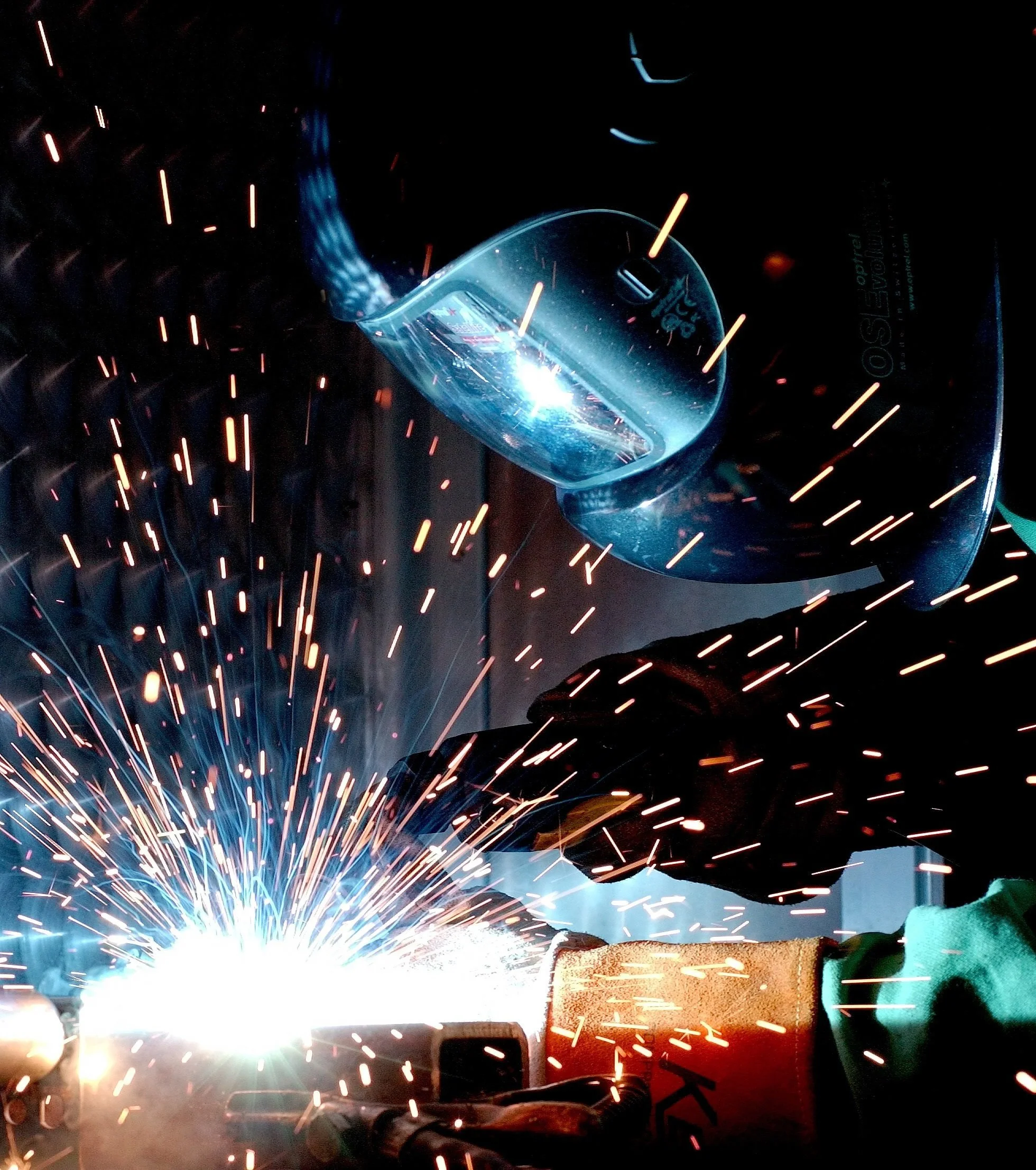 A person welding metal, with sparks flying in all directions and a protective welding helmet hanging above.