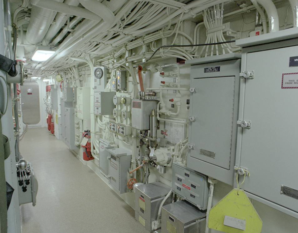 Interior of a long corridor with white pipes, electrical boxes, and control panels on the walls, likely in a ship or industrial facility.
