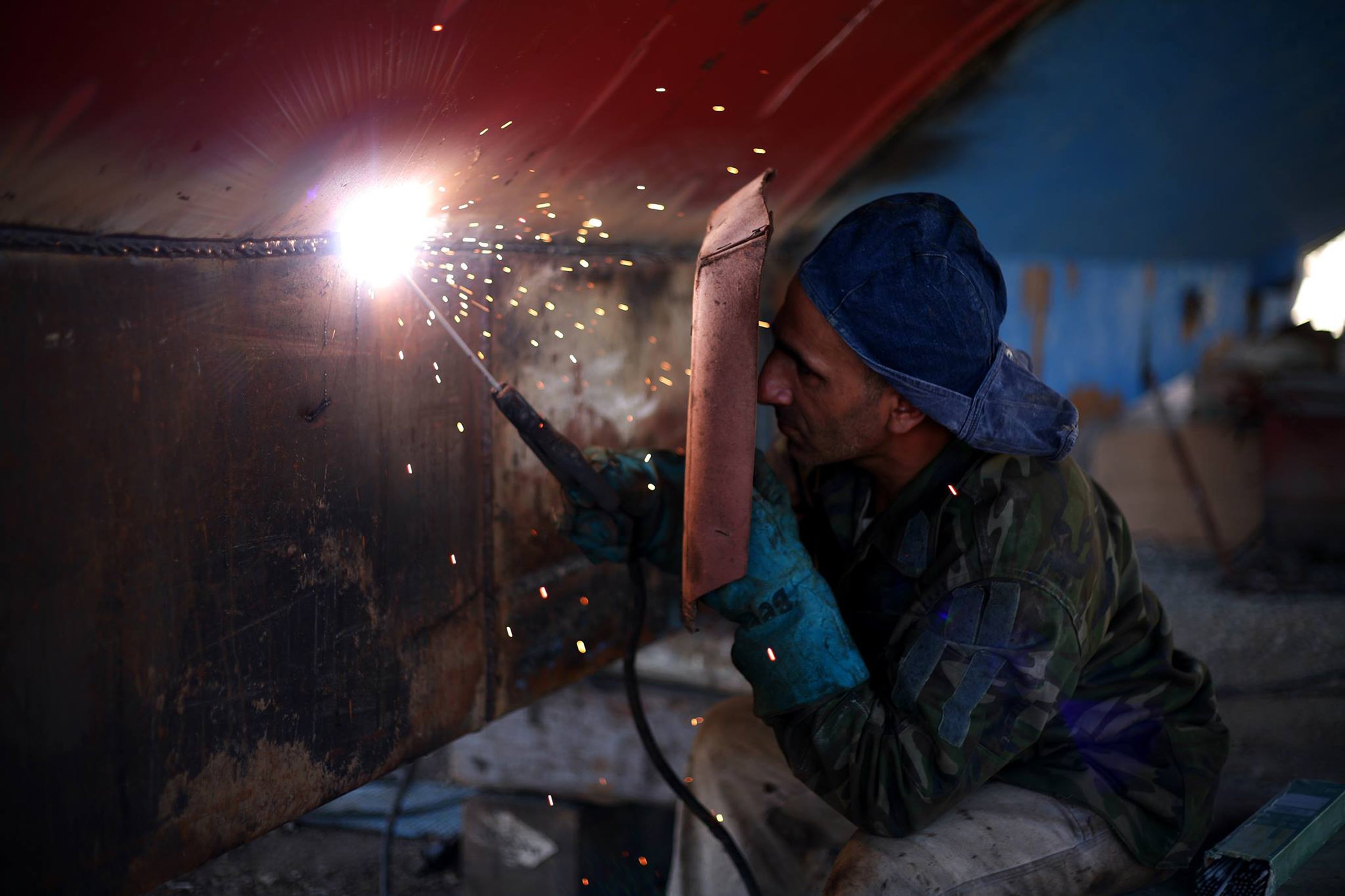 A worker welding a large metal structure with sparks flying in an industrial setting, wearing protective gloves, a denim hat, and camouflage shirt.
