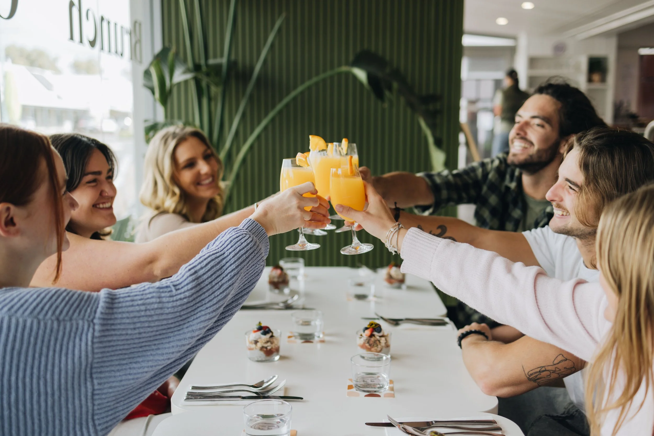 A group of friends celebrating with orange mimosas at a restaurant table, raising glasses in a toast, with desserts and utensils on the table.