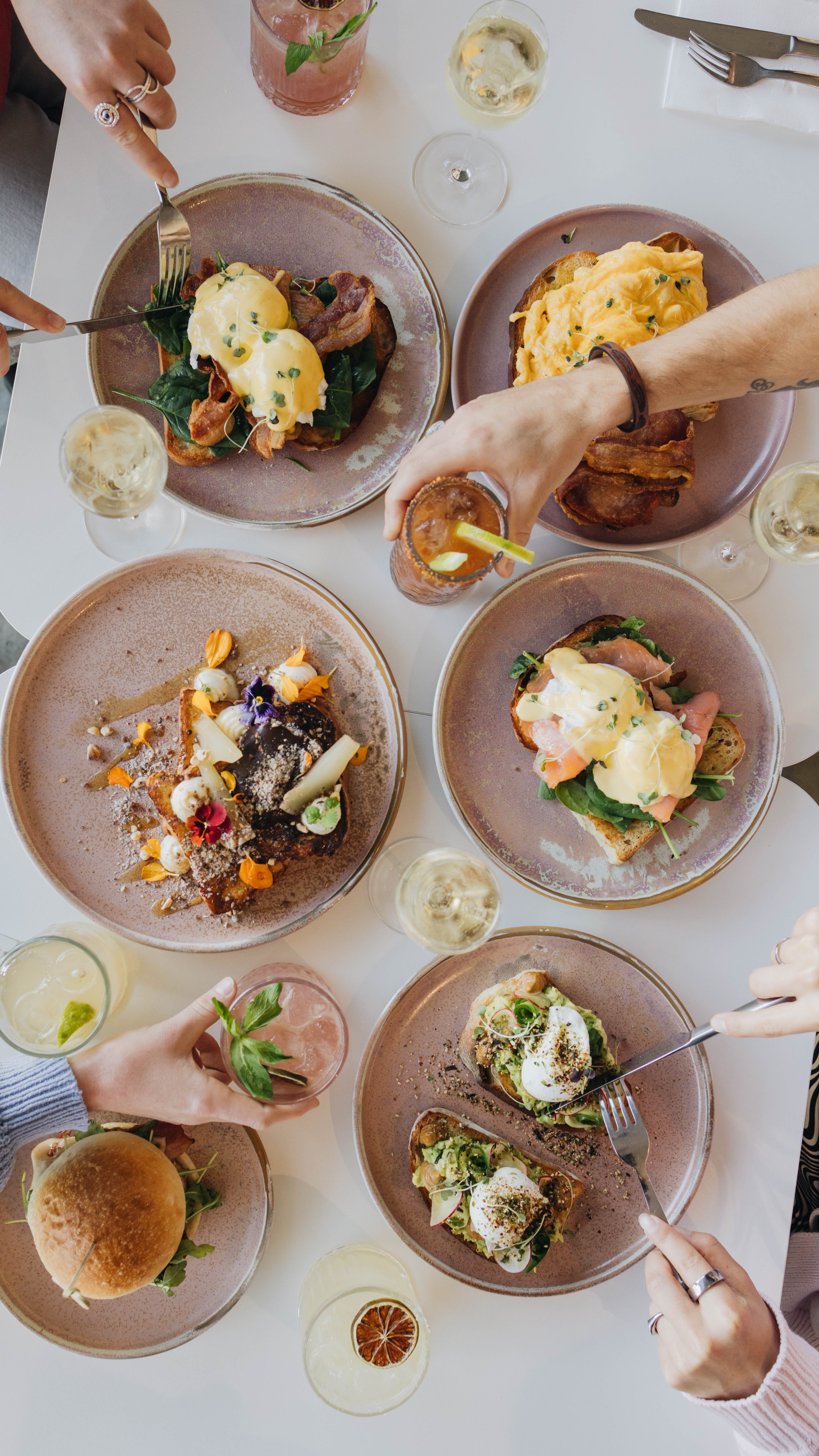 Overhead view of a table with various brunch dishes including eggs Benedict, a sandwich with smoked salmon and greens, a burger, and a dessert with edible flowers, along with glasses of mimosas and other drinks, with people reaching for the food.