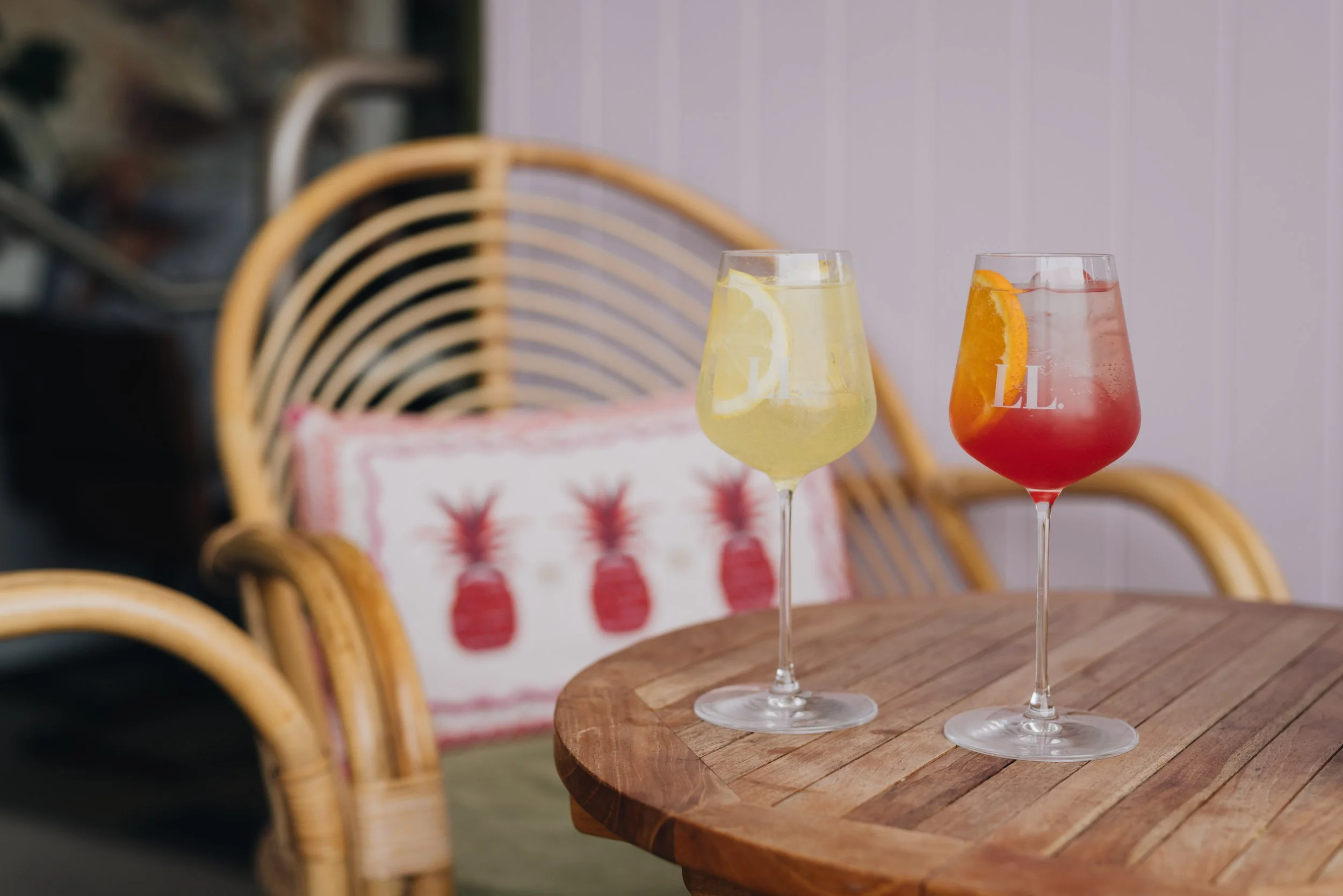 Two signature spritz cocktails in a room with wicker chairs and a pillow with pineapples.