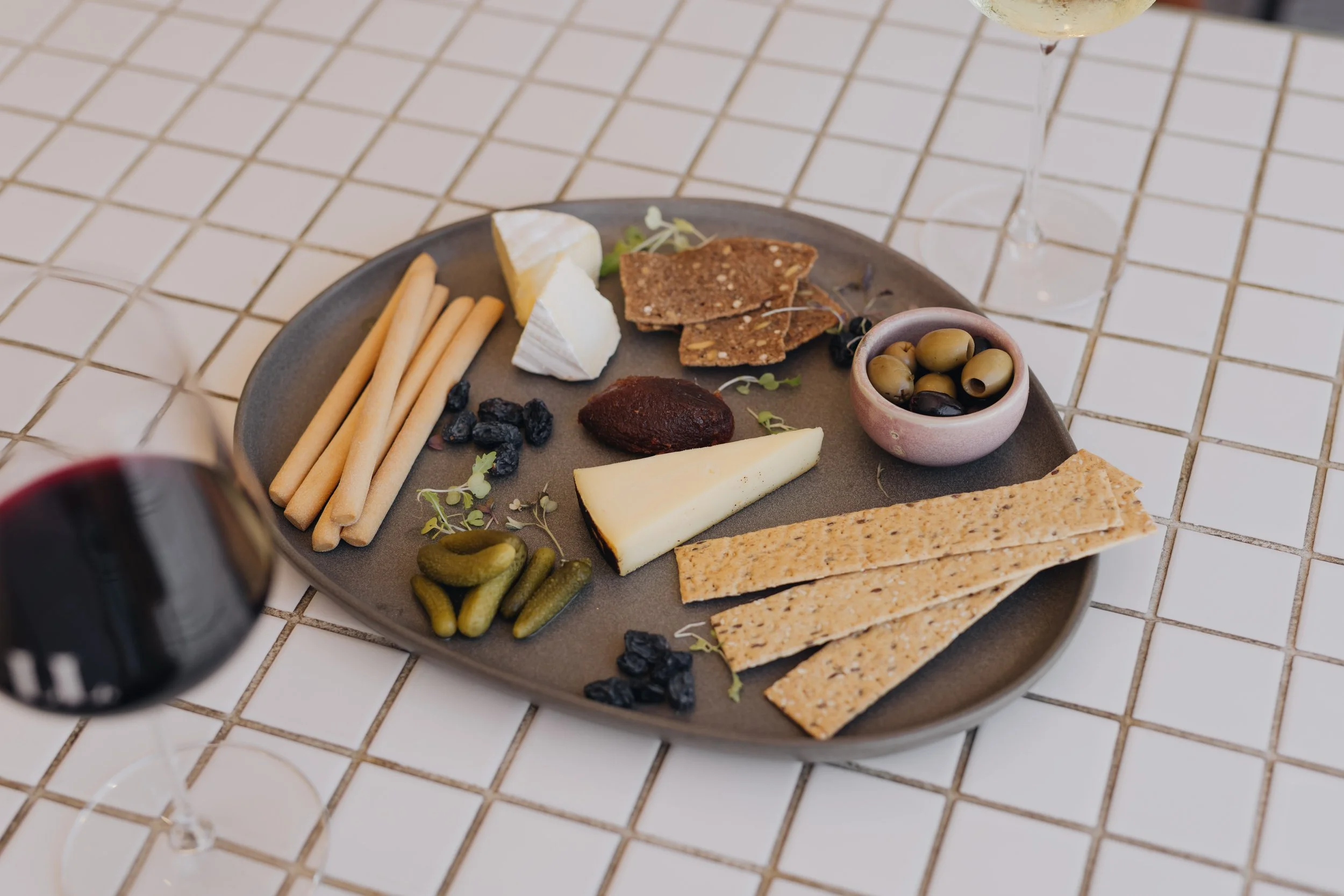 A gray oval plate with assorted cheeses, crackers, breadsticks, olives, pickles, and other snacks, with two glasses of wine on a white tiled table.