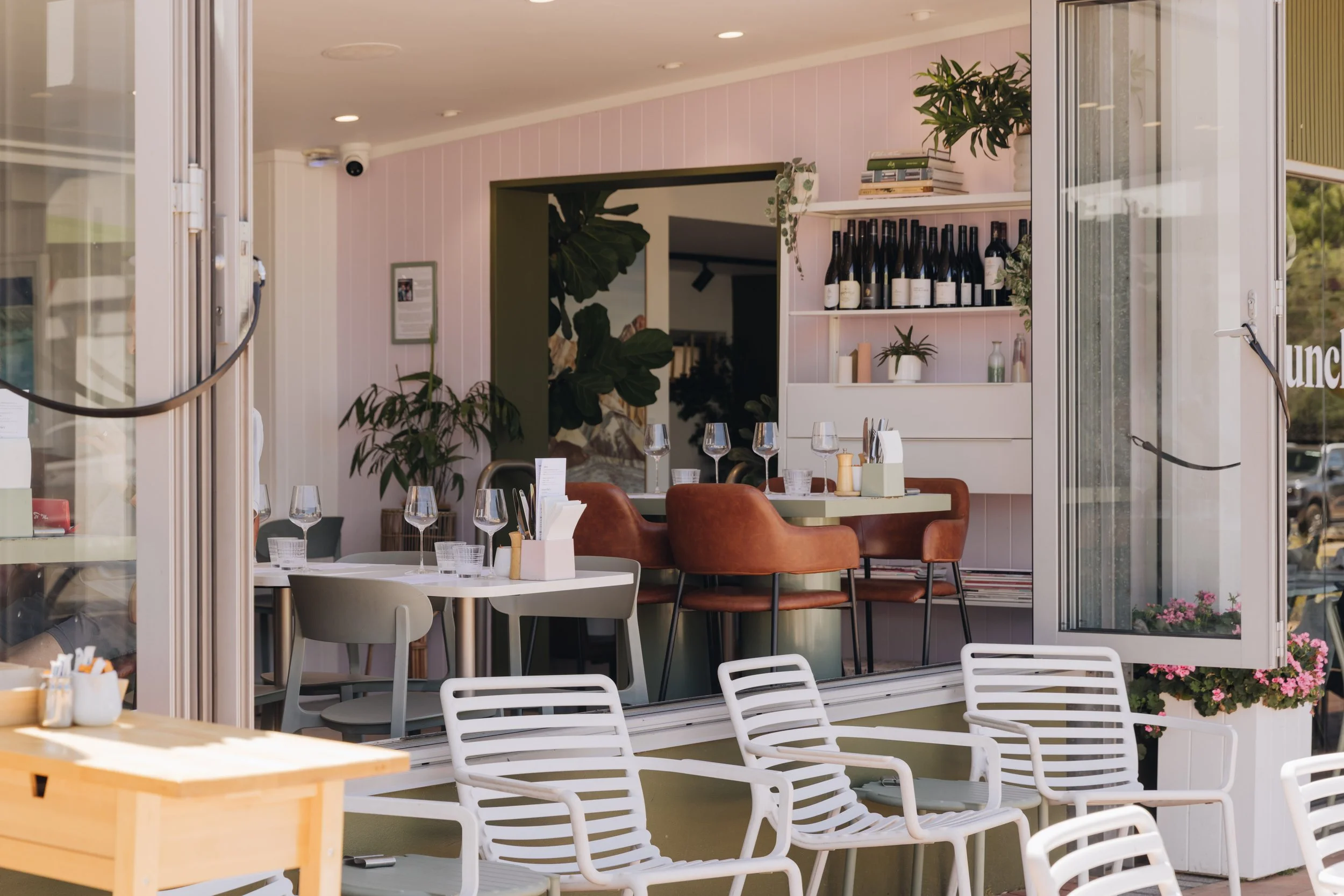 Interior of a bright, modern restaurant or cafe with a round dining table set with wine glasses and paper placemats, surrounded by brown chairs. There are shelves with wine bottles, books, and potted plants, and large windows with a view of outdoors,