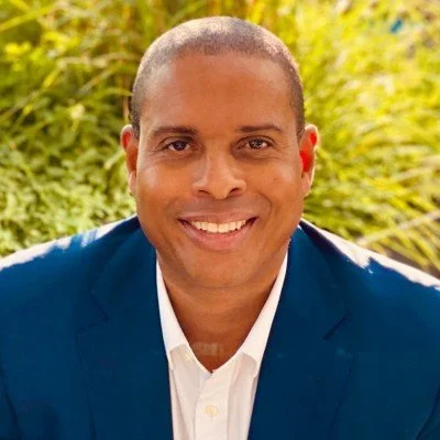 Portrait of a smiling man with short hair, wearing a white shirt and blue blazer, outdoors with green foliage in the background.