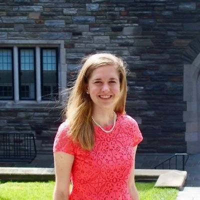 Young woman with long blonde hair smiling outdoors in front of a stone building, wearing a pink lace dress and a pearl necklace.