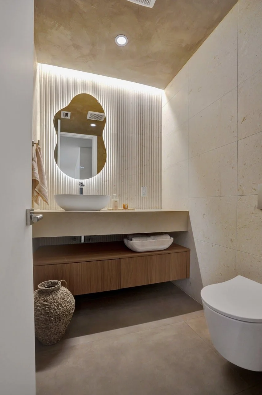 Modern bathroom with a vessel sink, an oval mirror, textured wall panel, and a floating wooden cabinet. Light-colored walls and floor, with a woven basket on the floor next to the cabinet.