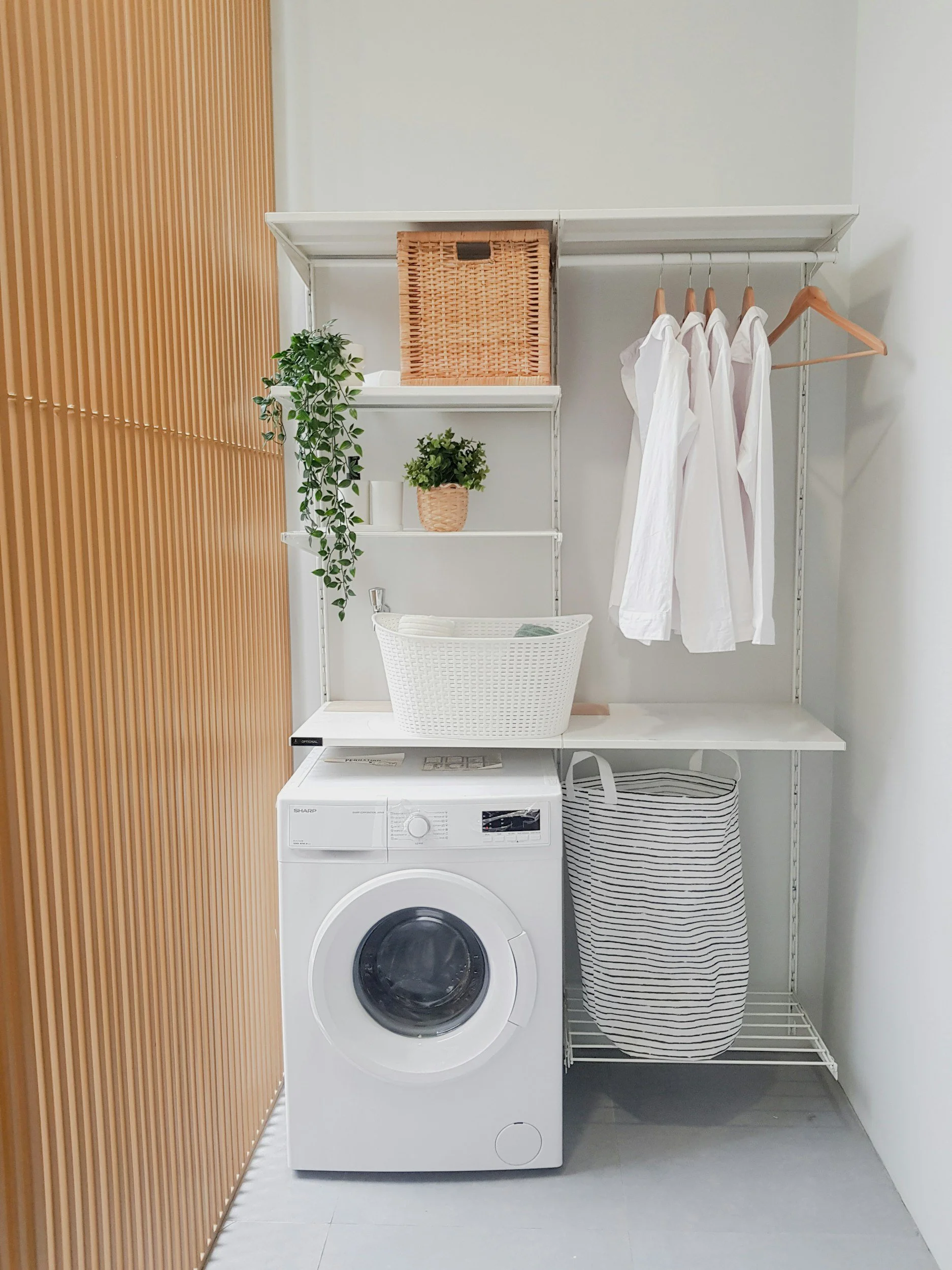 A laundry room with a white washing machine, storage shelves with plants and baskets, and a striped fabric laundry hamper.