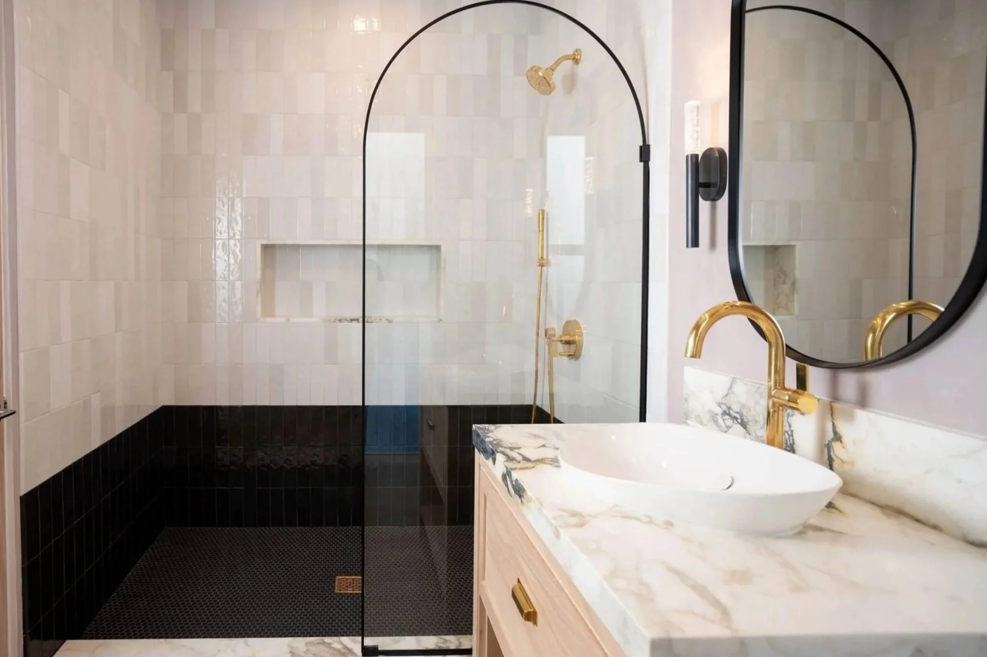 Modern bathroom with a white marble vanity, black accents, gold fixtures, and a glass shower with beige and black tile walls.