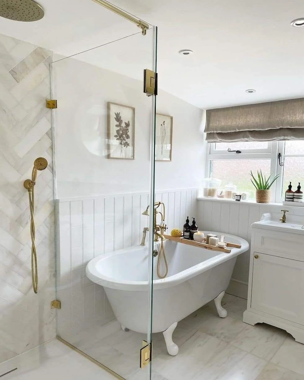 Bathroom with a clawfoot bathtub, gold fixtures, a glass shower enclosure, and a window with a beige valance. Decor includes framed wall art, plants, and toiletries.