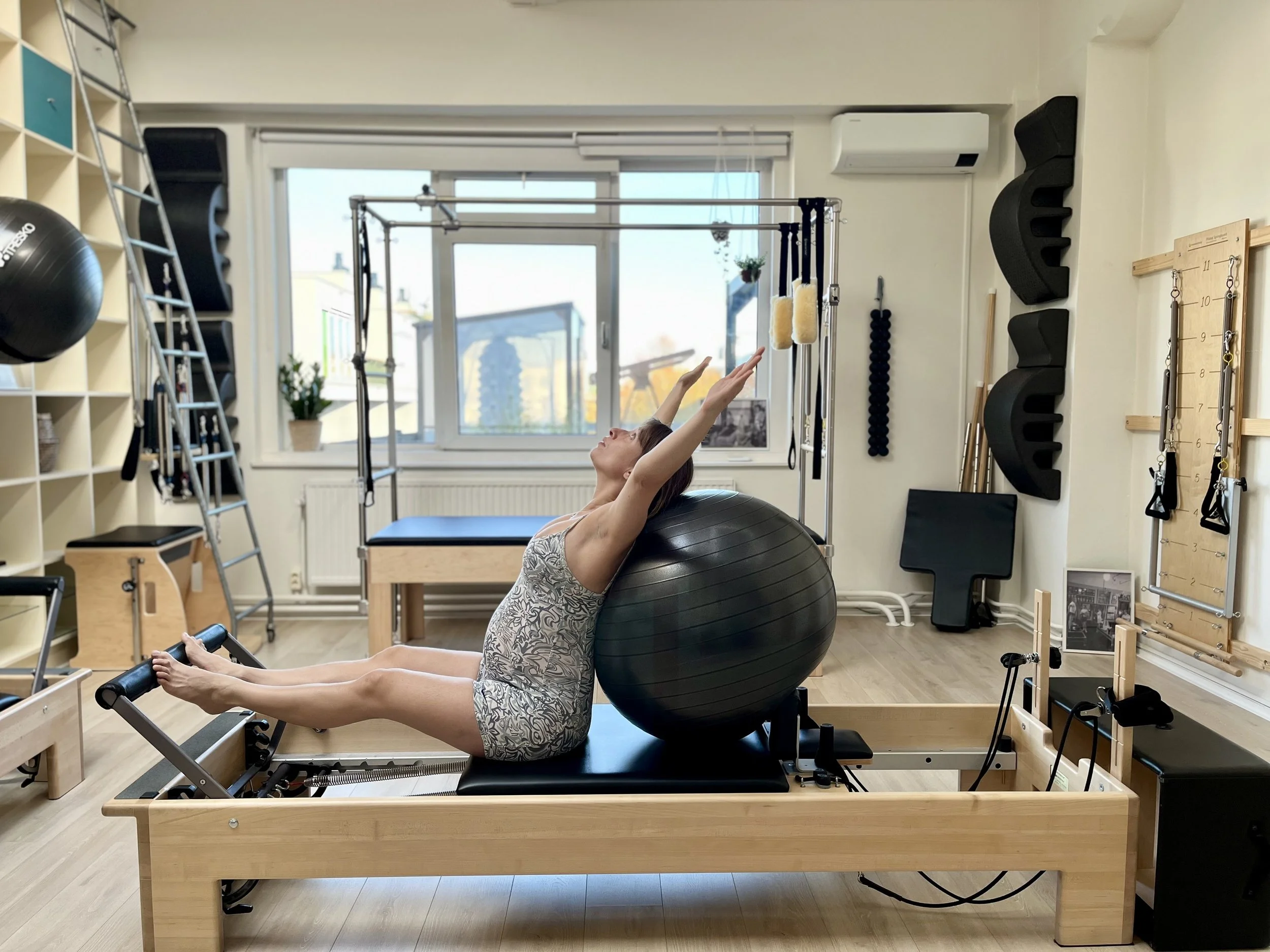 A woman doing Pilates on a reformer machine in a fitness studio, with her arms extended above her head and a large exercise ball supporting her back.