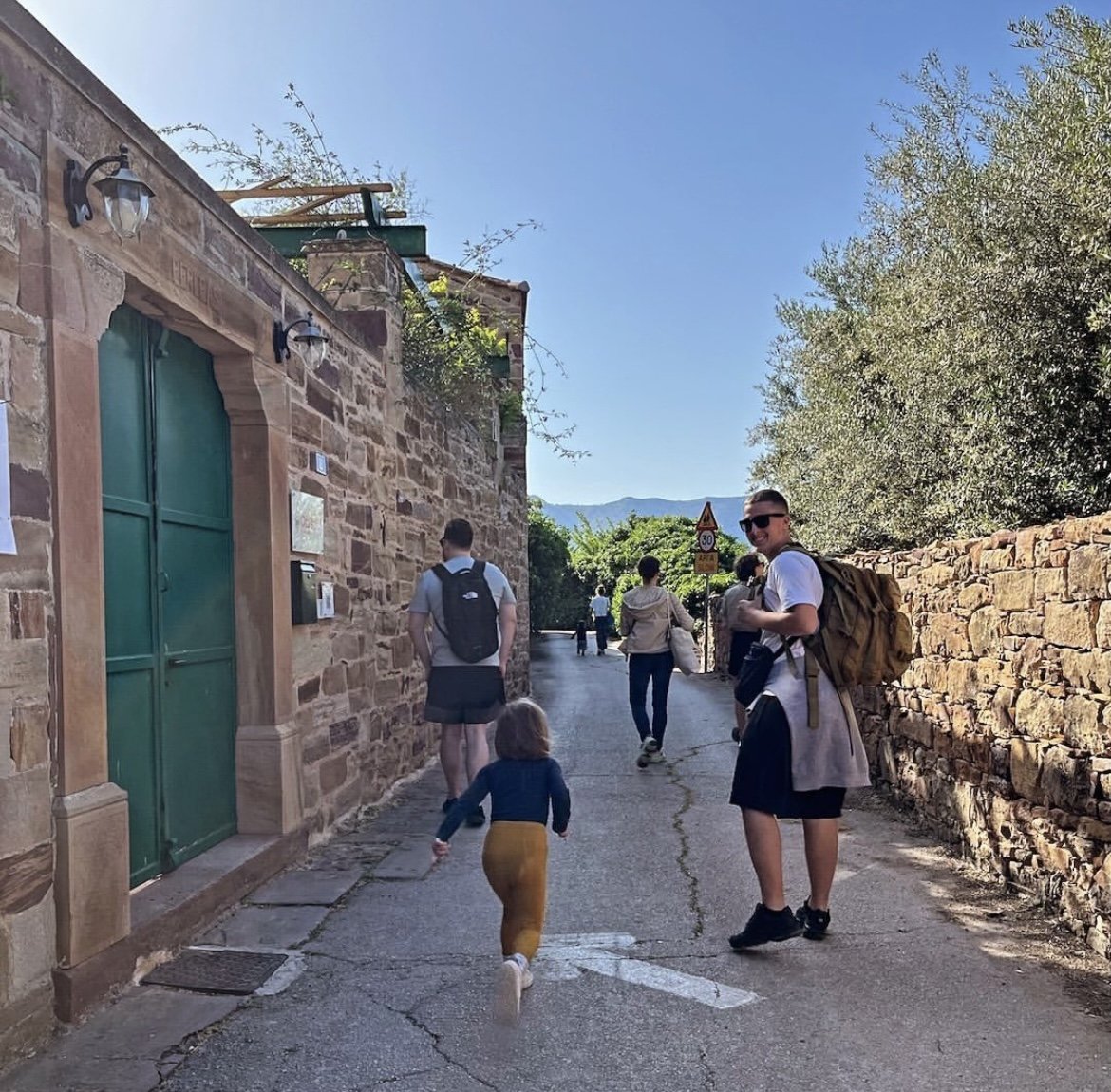 People walking down a narrow stone-paved street lined with stone walls and greenery, under a clear blue sky.