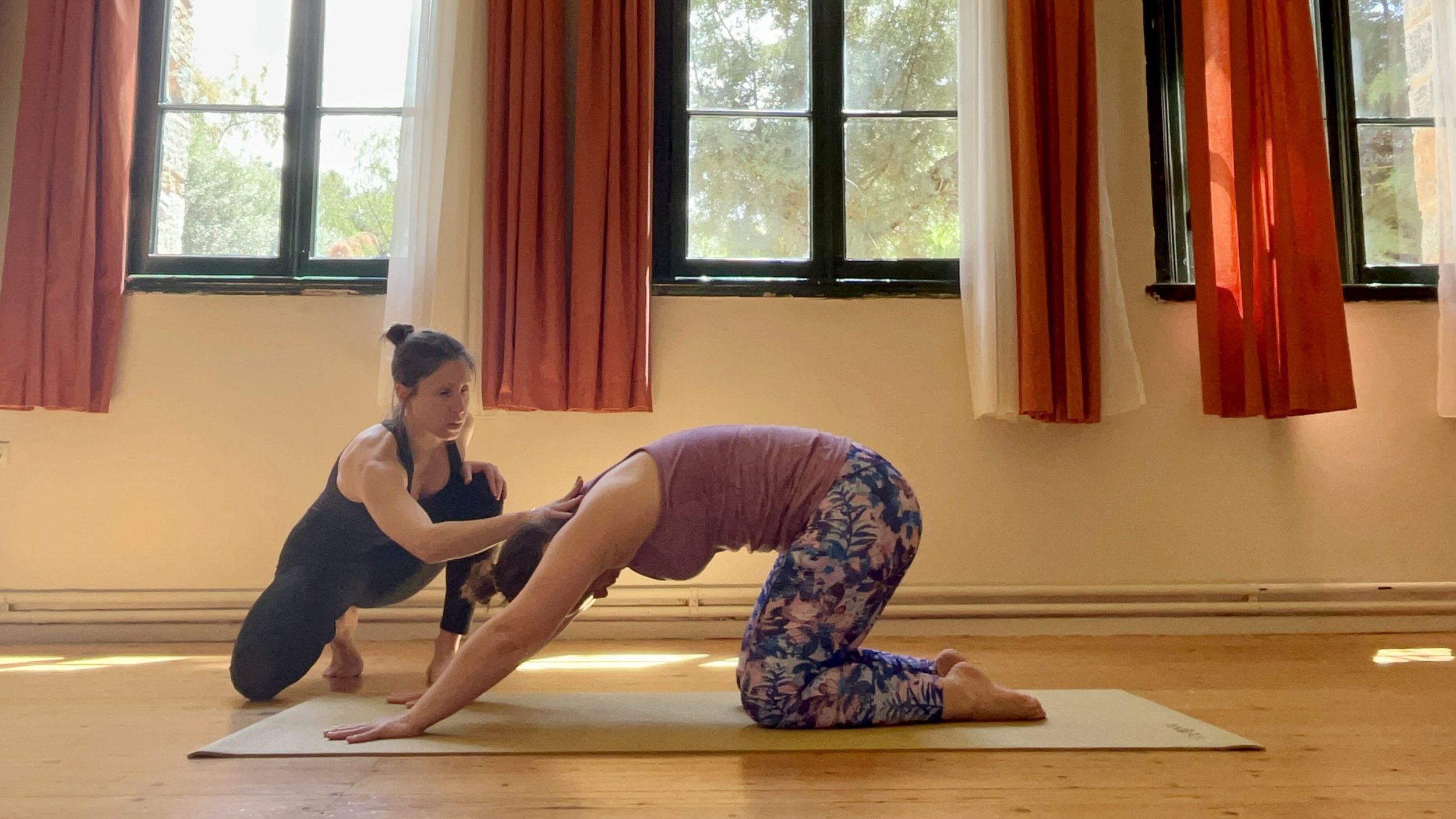 Two women practicing mat pilates indoors on a wooden floor, with windows and curtains in the background. One woman is in a child's pose, and the other is assisting or guiding her.
