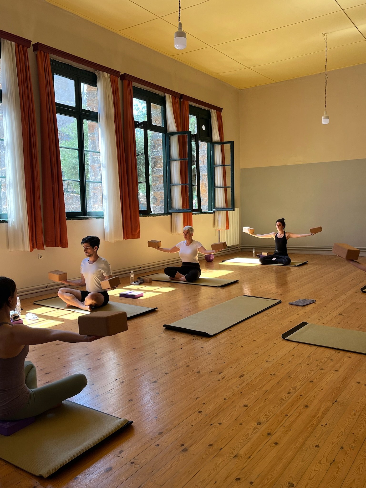 People practicing yoga in a well-lit room with wooden floors, large windows, and yoga mats. They are using yoga blocks and sitting with legs crossed.