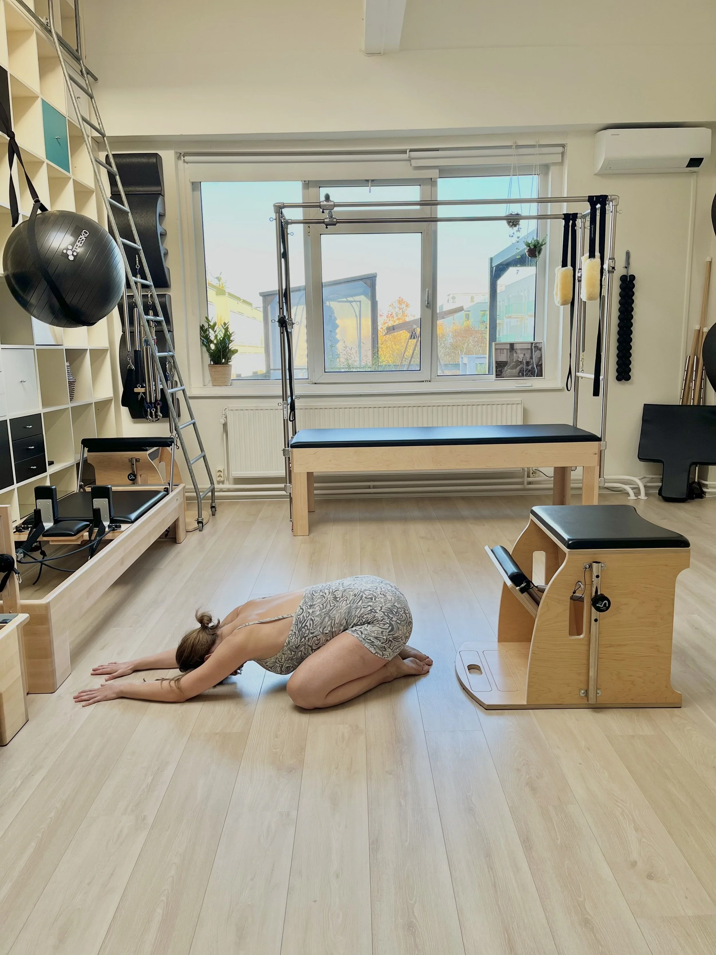A woman practicing yoga in a fitness studio with wooden floors, exercise equipment, and large windows showing trees outside.