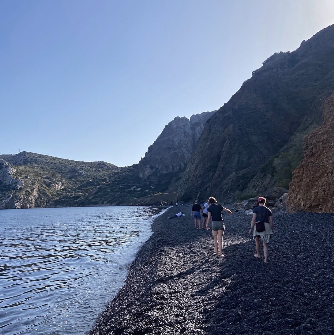 People walking along a rocky beach by the sea with mountainous cliffs in the background.
