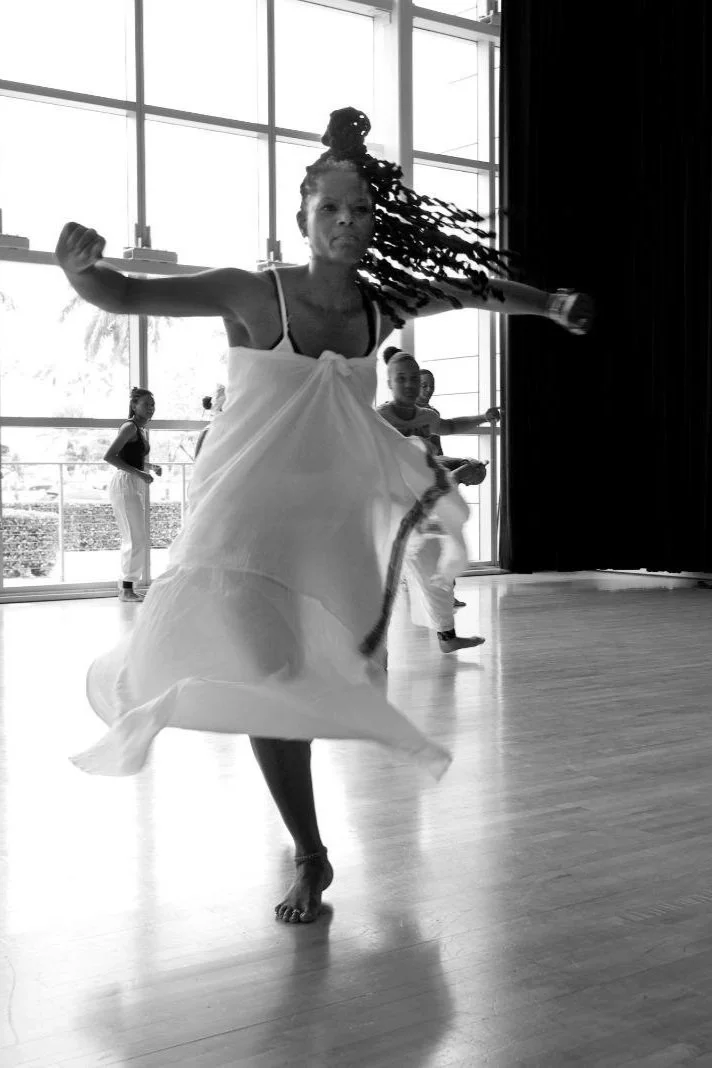 A woman dancing barefoot in a flowing dress in front of a large window with children behind her, in a spacious room.