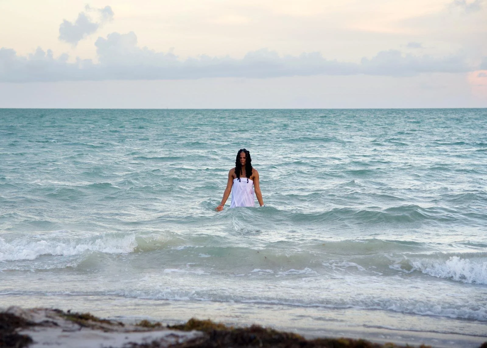 Woman in a white dress standing in the ocean, with waves around her, under a cloudy sky.