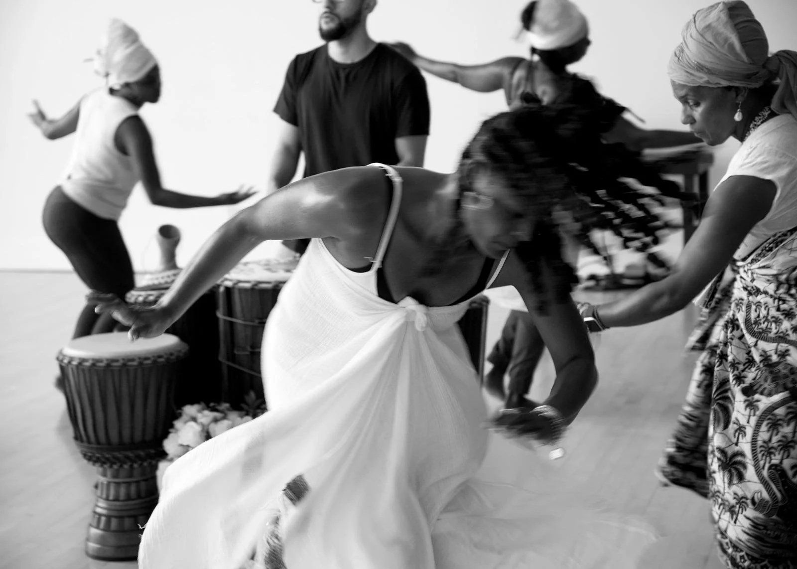A woman in a white dress bends forward as she is guided by a woman in patterned clothing. Other women in headscarves and turbans, and a man in a black shirt, are in the background, engaged in a cultural dance or celebration with drums present.
