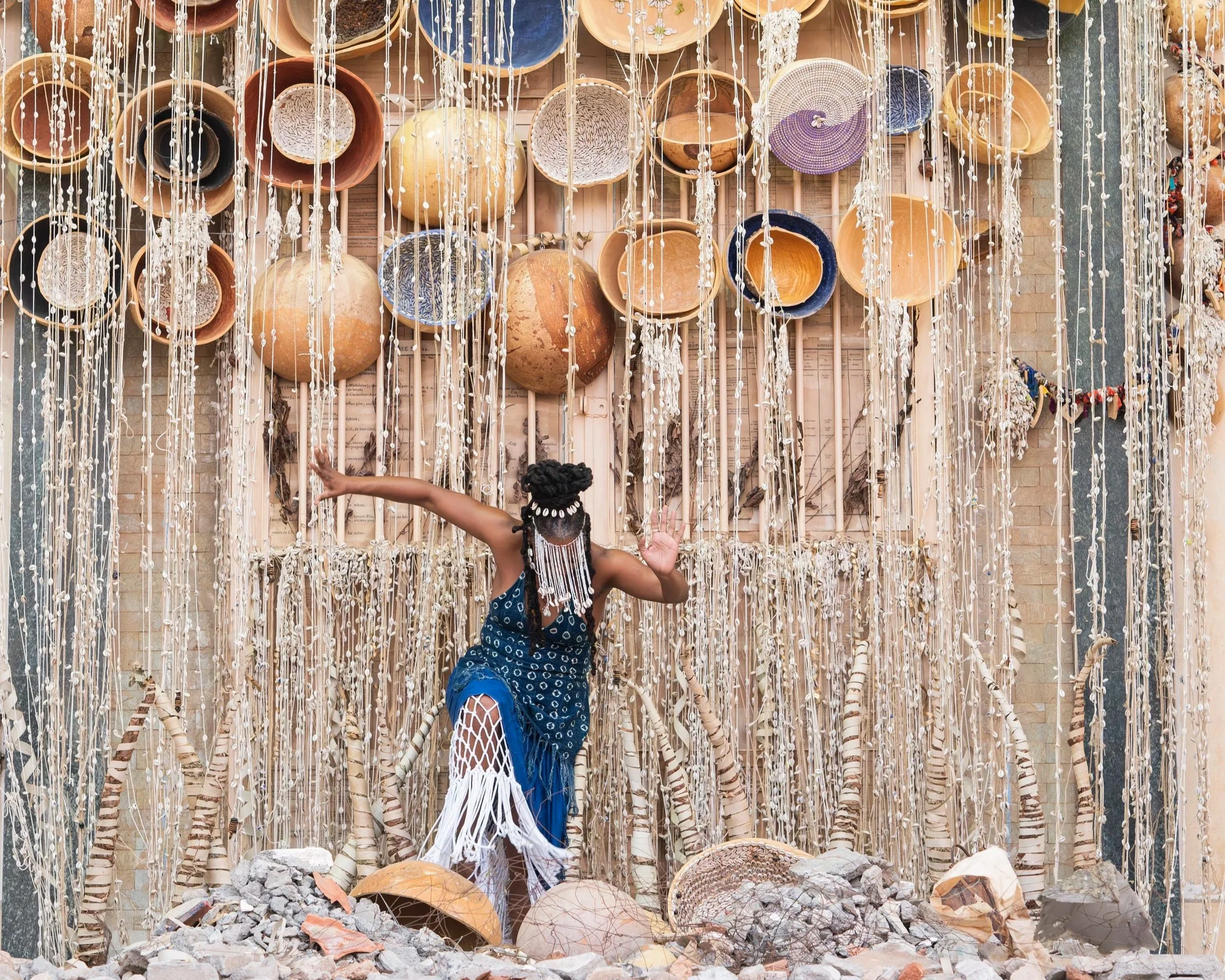 A person with braided hair and wearing a blue patterned dress is reaching out towards a wall decorated with hanging strings of shells, with terracotta pots and rocks at the ground, and a brick wall in the background.