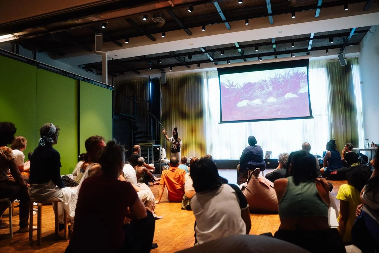 A woman in a patterned shirt stands on a stage pointing at a large screen displaying an outdoor landscape scene with rocks and greenery. The audience sits on chairs, stools, and the floor watching her.