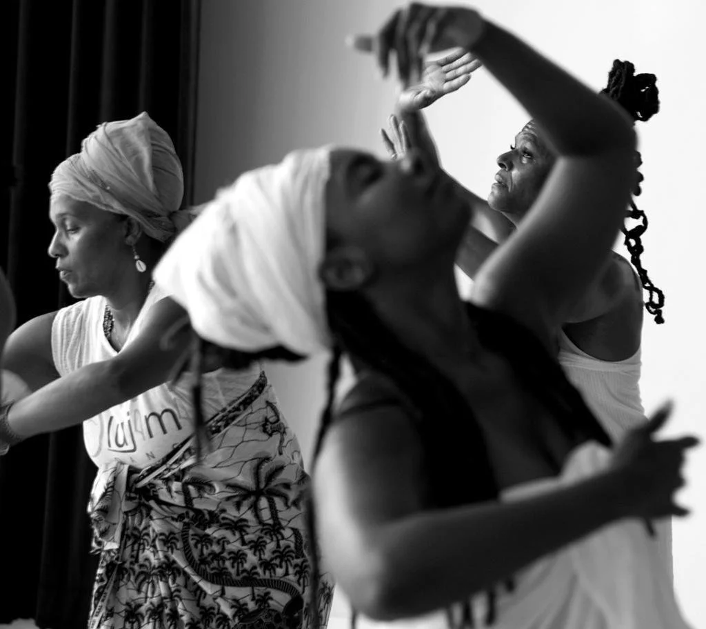 Three women dressed in traditional African attire, dancing or performing with expressive movements, one with a headwrap, in a room with a plain or white background.