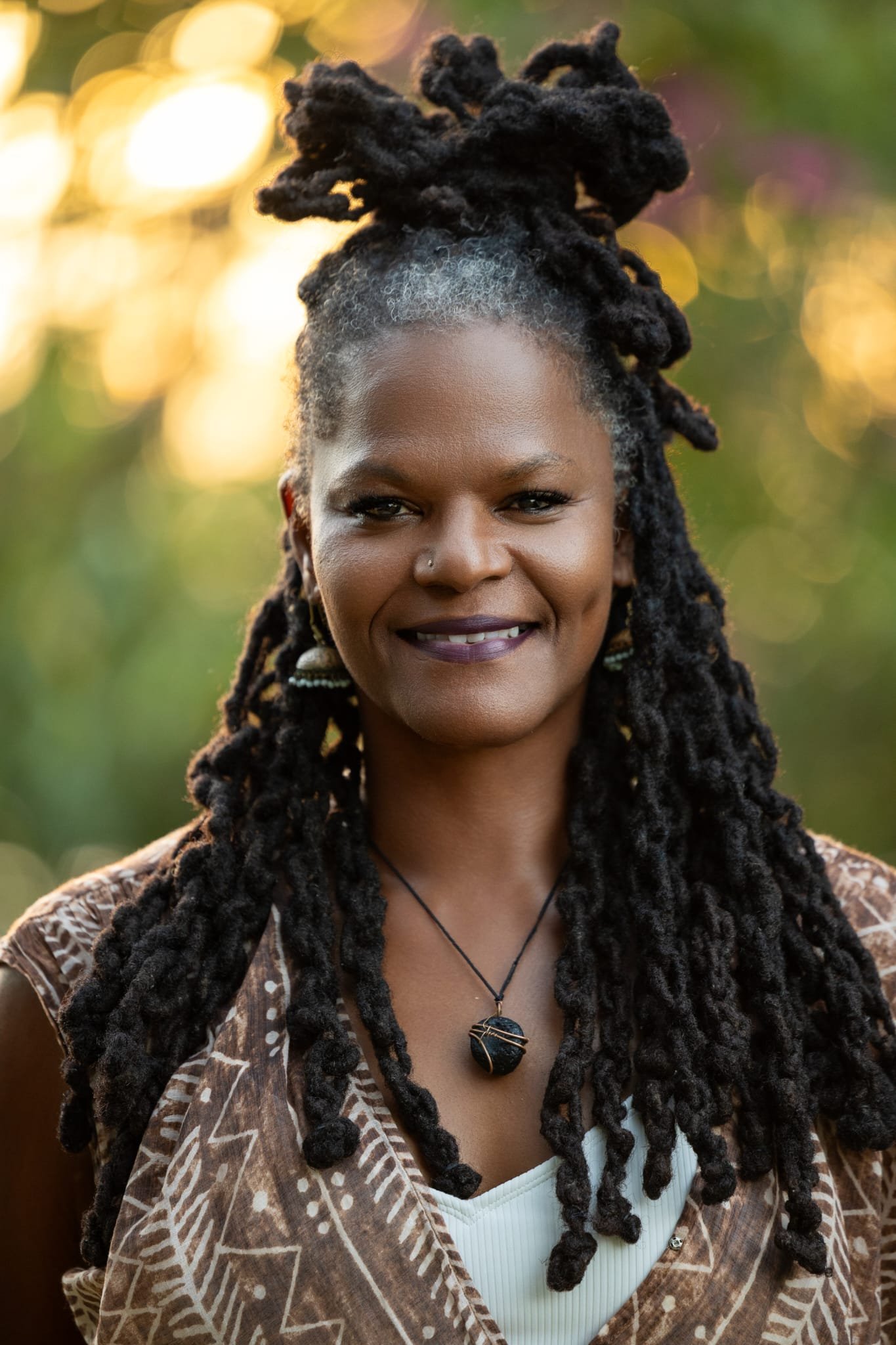 A woman with dark skin and long black locs smiling outdoors, wearing a patterned brown jacket, a white top, and a black pendant necklace, with a background of bright yellow and green bokeh lights.