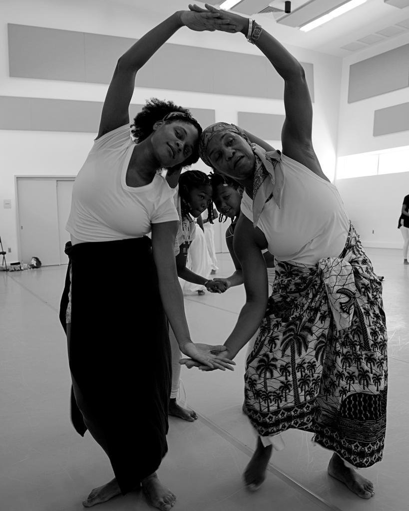 A group of people practicing yoga or dance together indoors, forming a connected pose with arms raised and hands joined.
