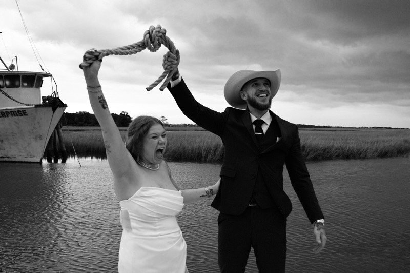 A bride and groom celebrate joyfully by a river, holding a rope with a docked boat and marshland in the background. The bride wears a strapless dress and the groom is in a suit with a cowboy hat.