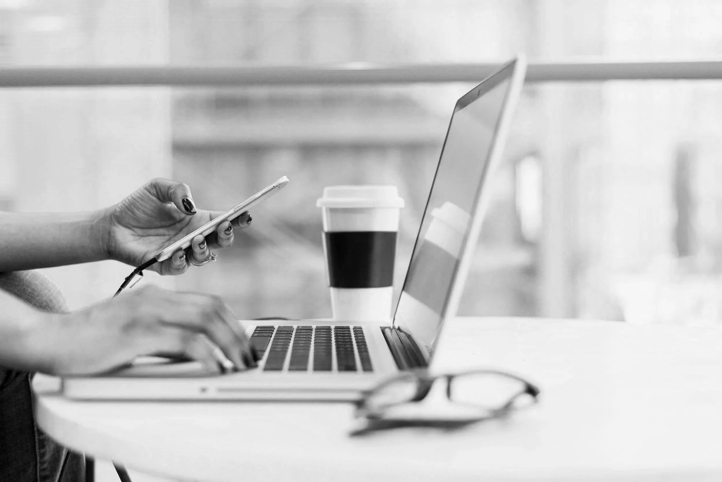 A person with manicured nails is using a smartphone while working on a laptop at a table, with a coffee cup nearby.
