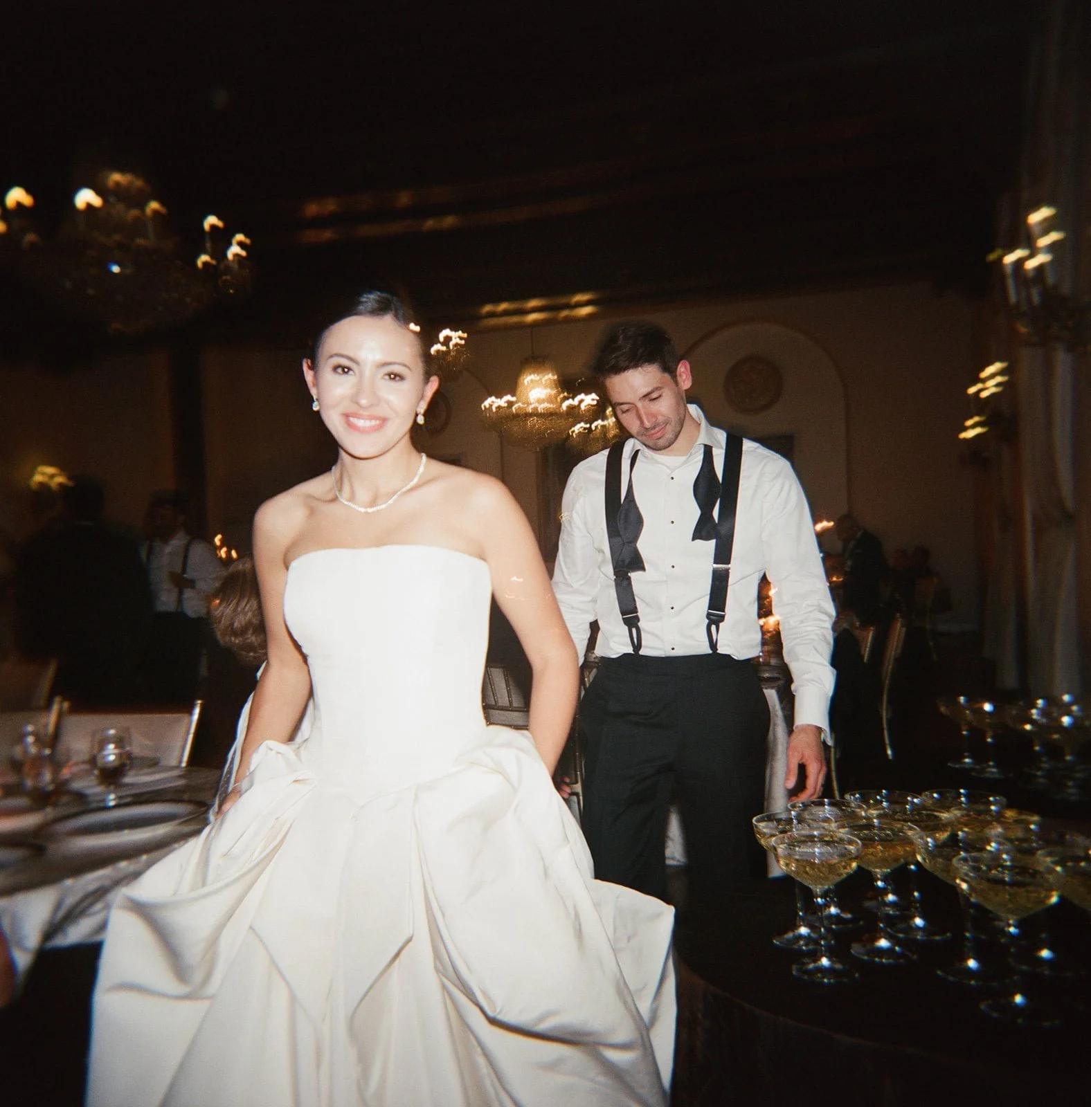 A smiling bride in a white strapless wedding gown and pearl necklace standing in a decorated reception hall, with a man in a white shirt with suspenders behind her, and glasses of champagne on a table nearby at the St. Regis Hotel in Washington DC.