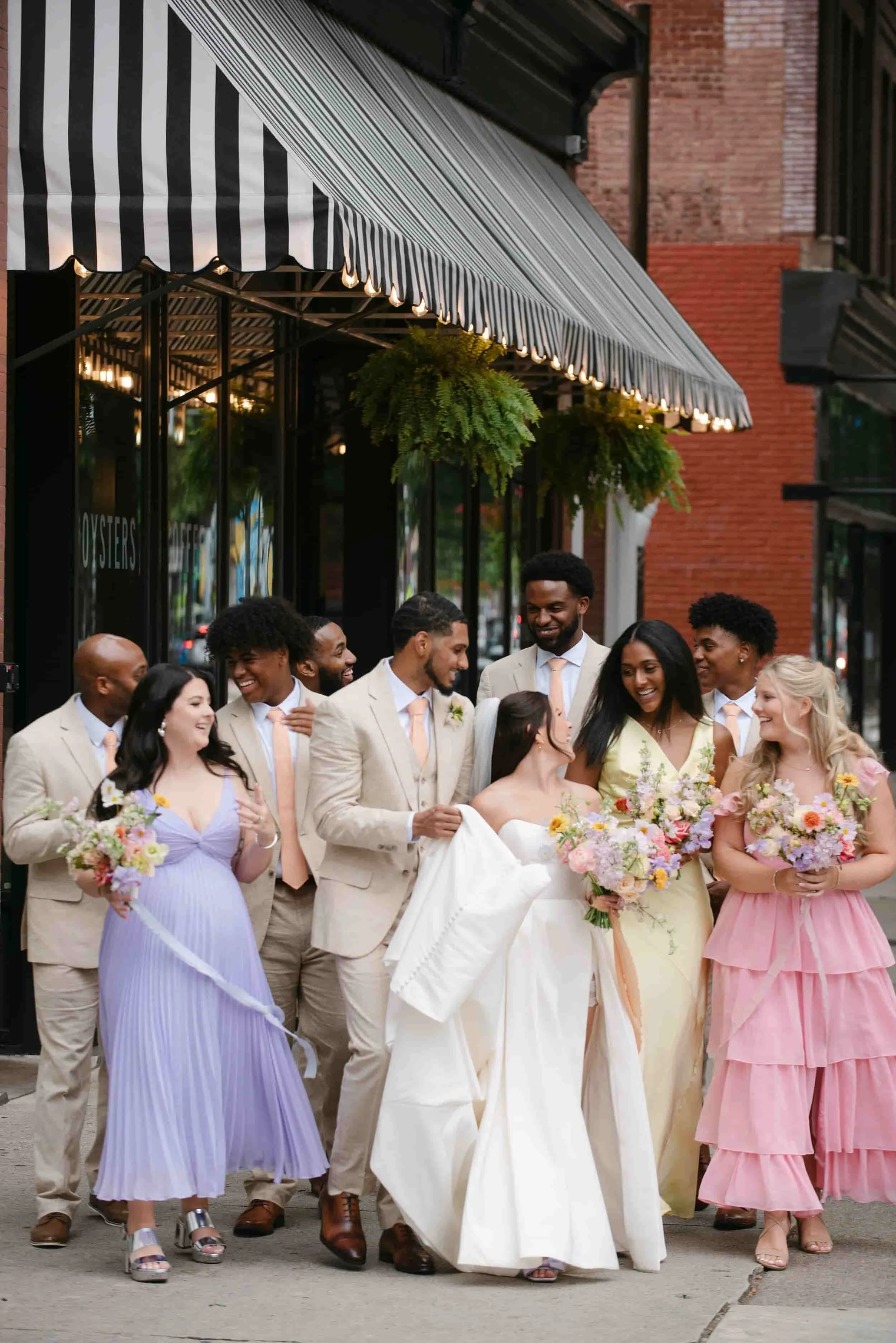 Group of people dressed in formal attire celebrating on city sidewalk, holding flowers, outside a storefront with black and white striped awning at Common House Richmond