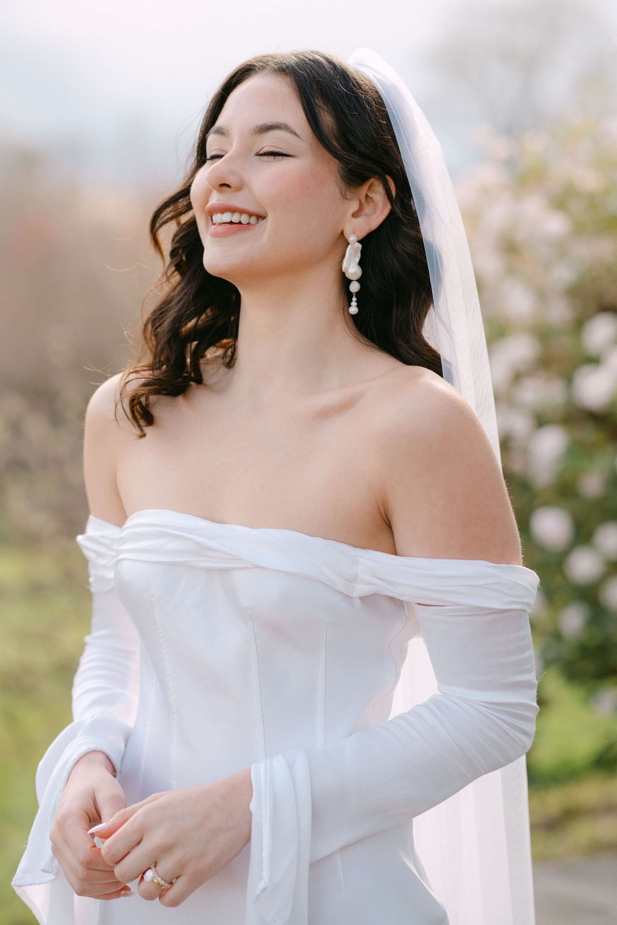 A smiling woman in a white off-shoulder wedding dress with a veil and pearl earrings outdoors.