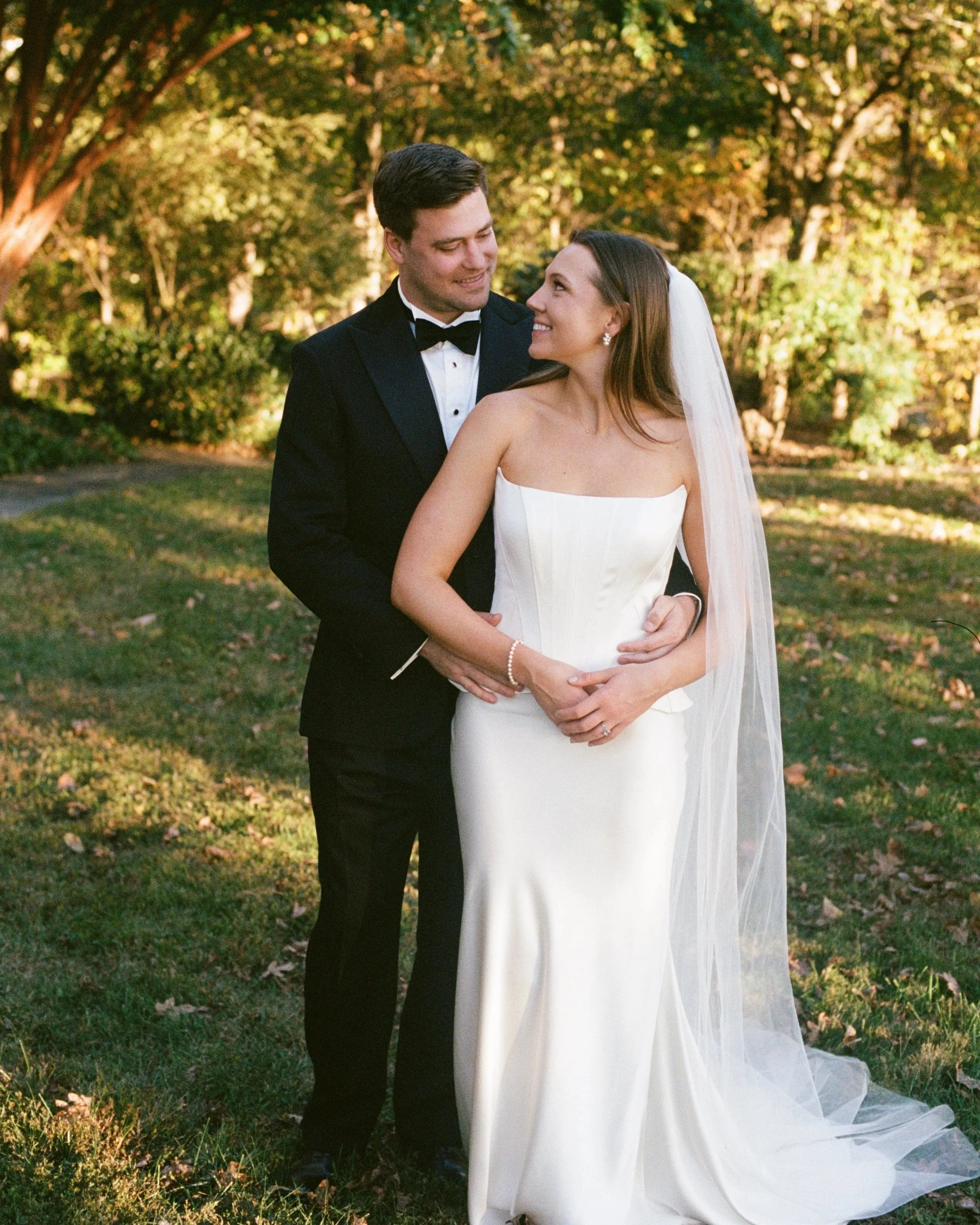 A bride and groom standing outdoors on a grassy area with trees in the background, smiling at each other. The groom is wearing a black tuxedo with a bow tie, and the bride is dressed in a strapless white wedding gown with a long veil at Goodstone Inn