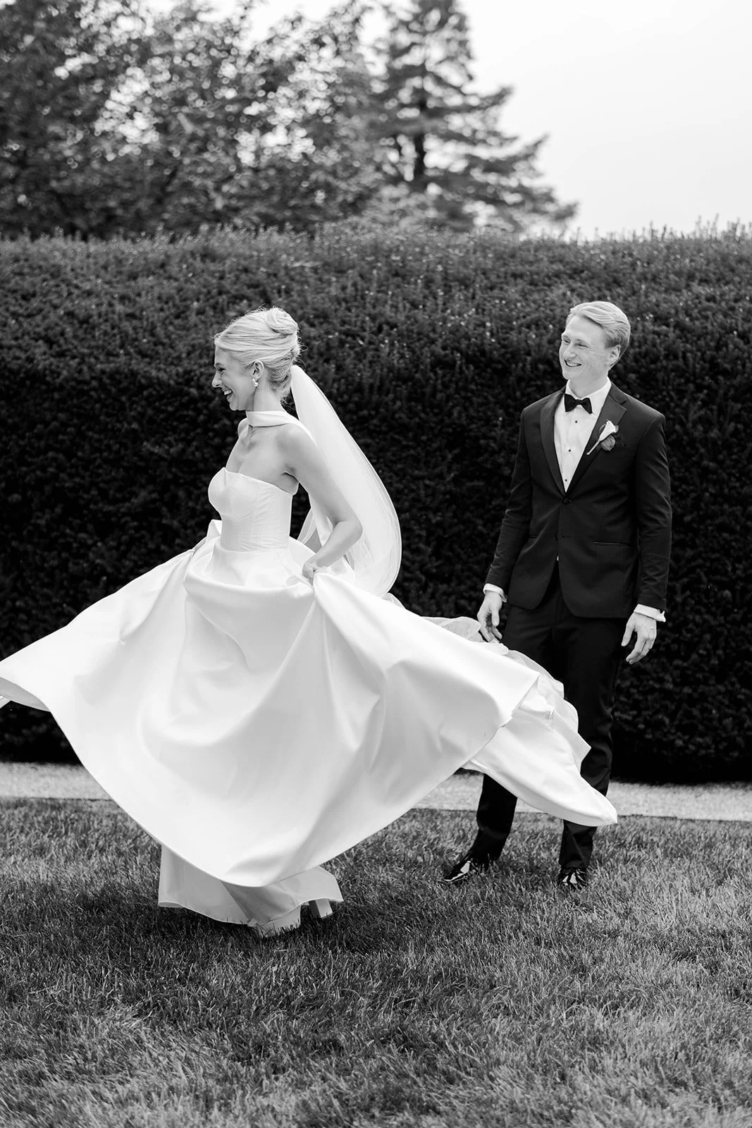 A bride in a wedding dress, smiling and spinning on a lawn, with a groom in a tuxedo watching her, against a backdrop of bushes and trees.