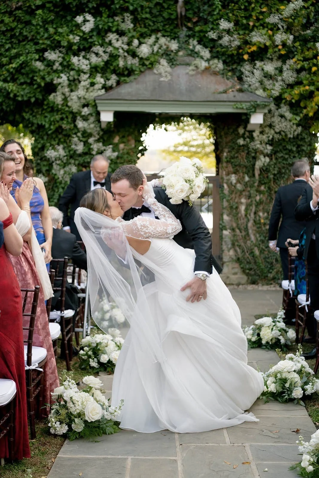 A newlywed couple shares a kiss during their wedding ceremony, with the groom dipping the bride. Guests are clapping and smiling, and the setting is decorated with white flowers and greenery at The Goodstone Inn in Middleburg VA