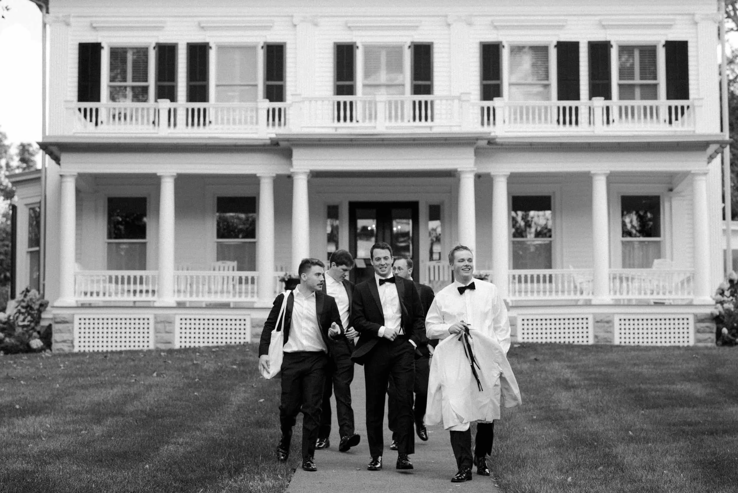 Black and white photo of a group of five men dressed in tuxedos, with one man carrying a white garment bag, walking outside in front of a large, colonial-style house with columns and a second-floor balcony.