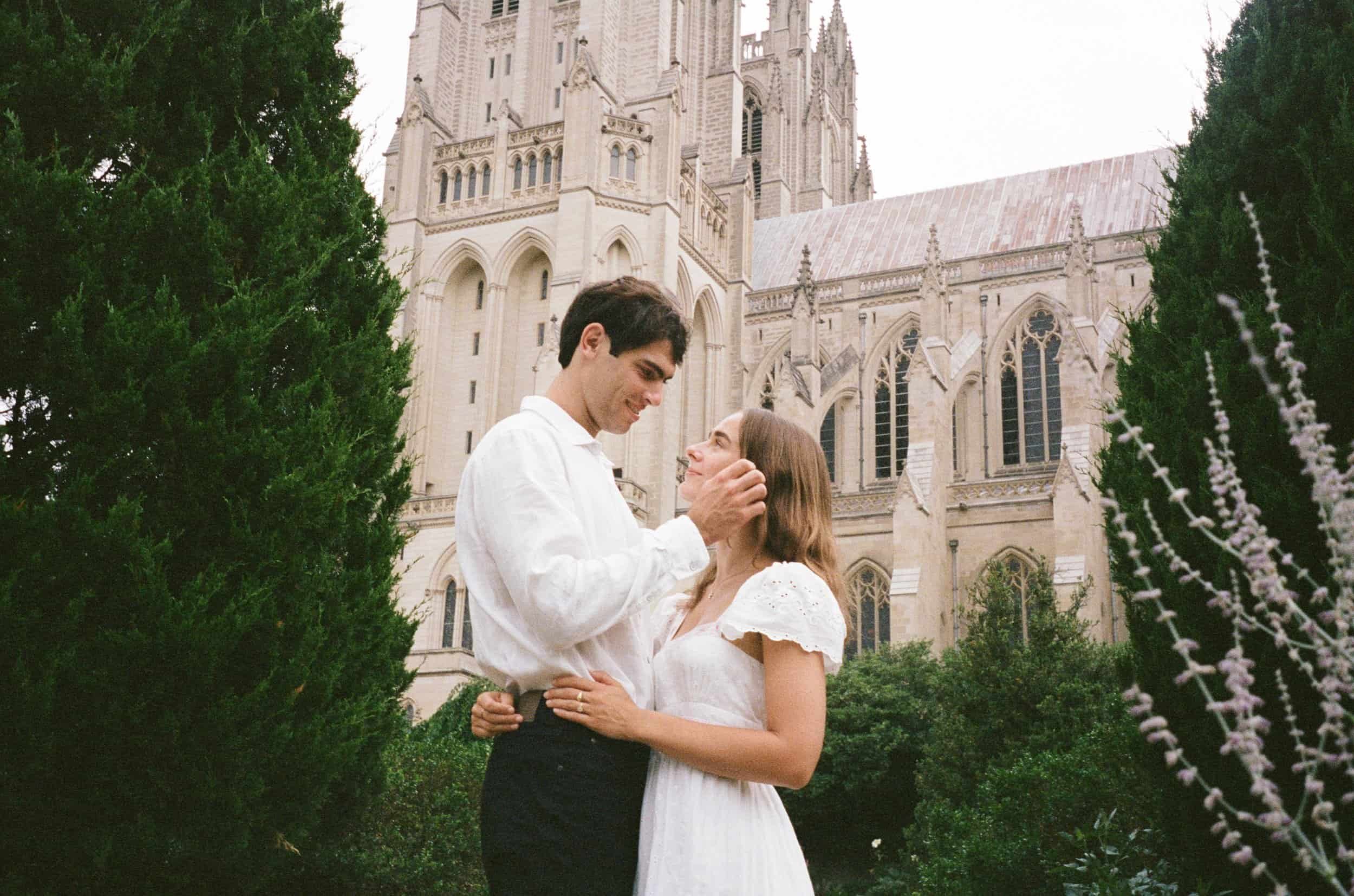 A couple standing close to each other outdoors in front of a large gothic-style cathedral. The man is holding the woman's face gently. The woman is smiling and looking up at the man. The woman is wearing a white dress with lace sleeves, and the man i