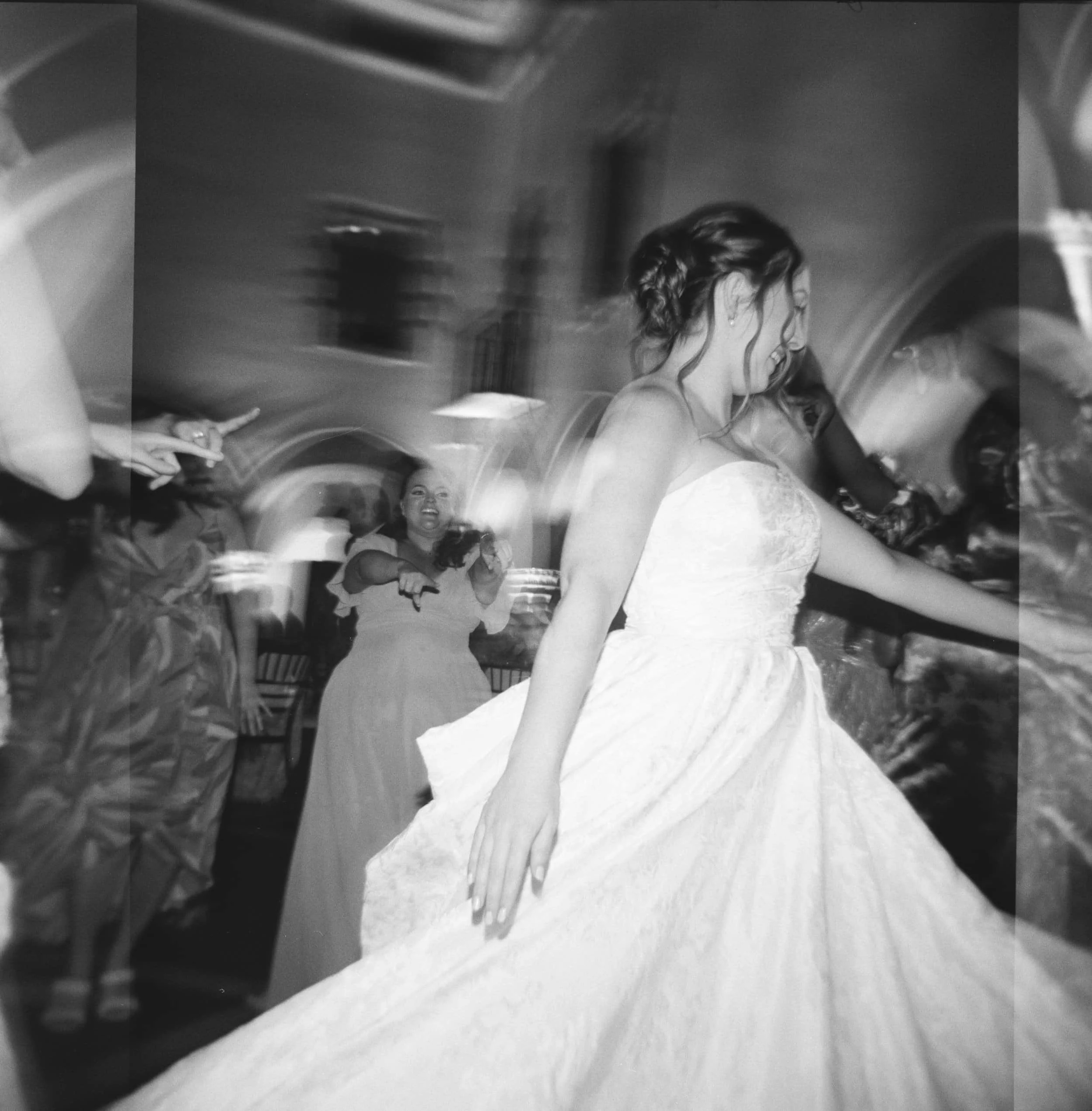 A woman in a strapless wedding dress spinning on a dance floor at a celebration, with guests dancing around her and one guest in the background smiling and pointing.