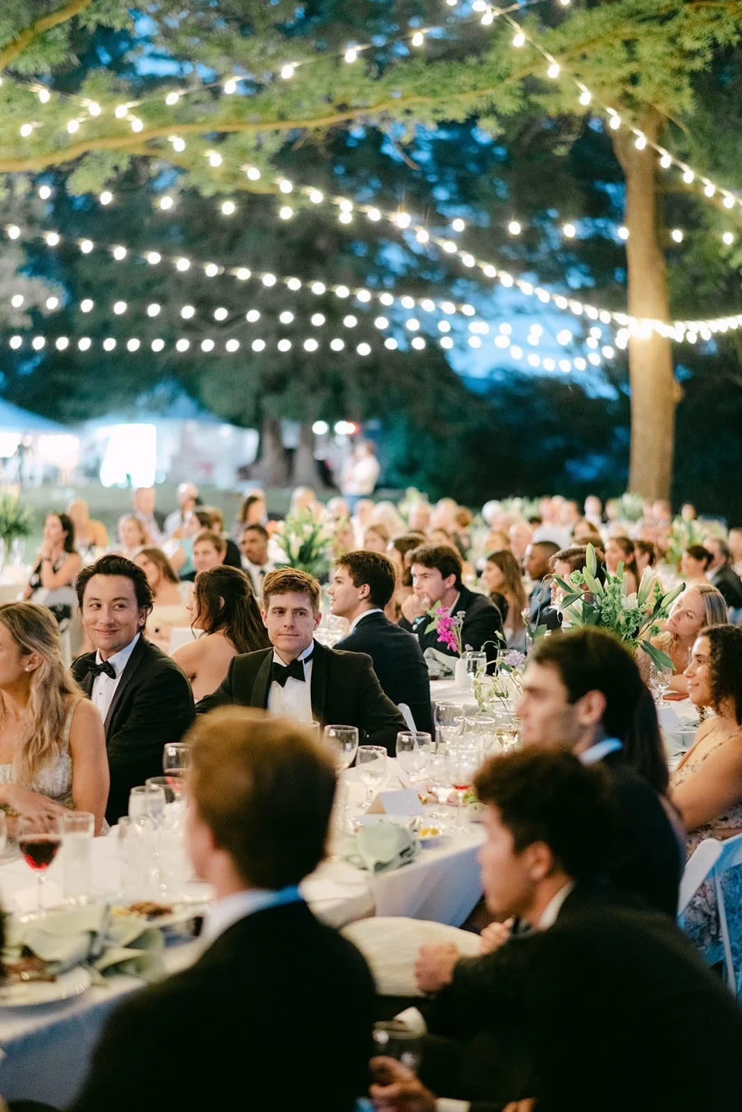 People dressed in formal attire attending an outdoor evening event under string lights, with tables decorated with flowers and food.