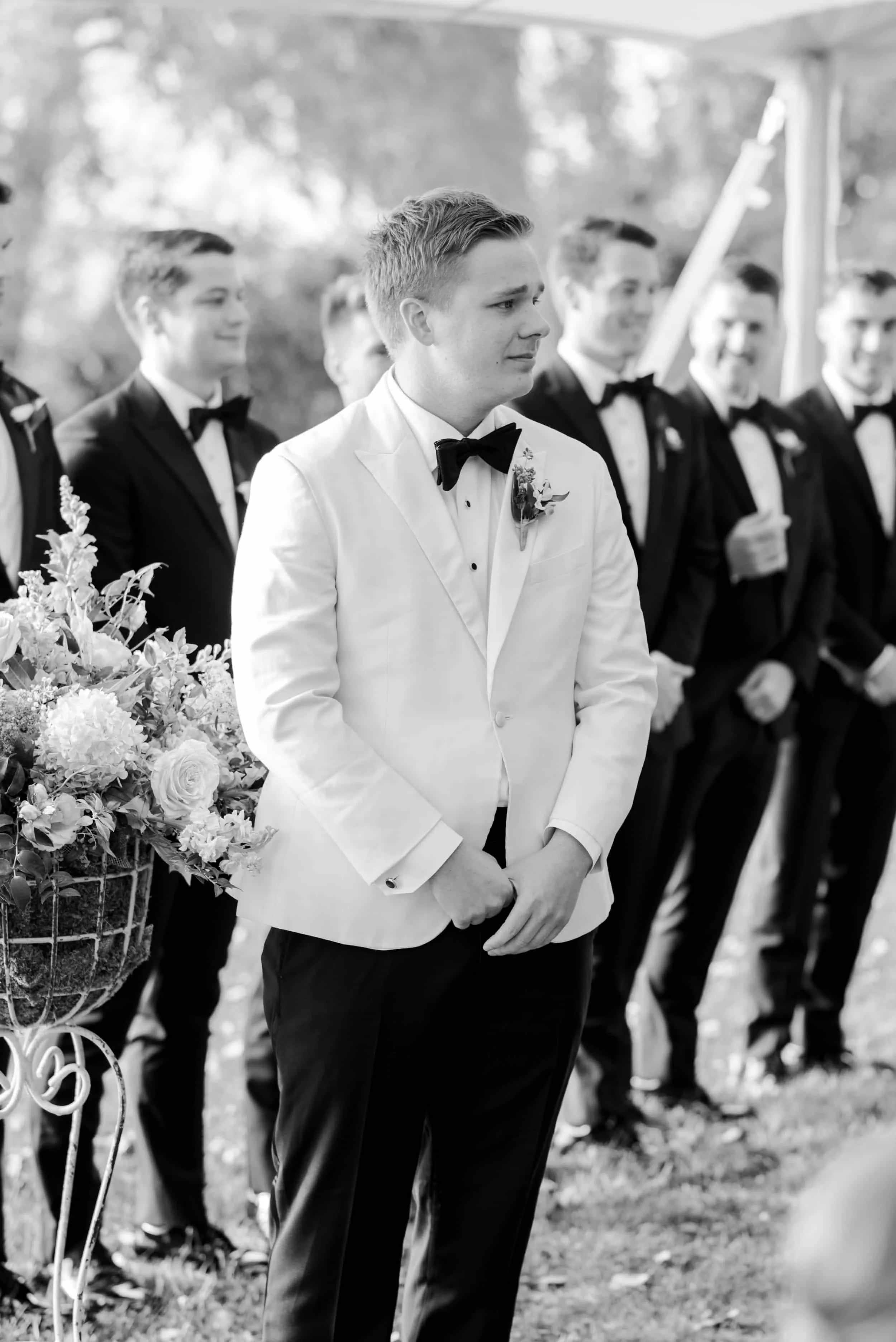 A groom in a white suit jacket and black bow tie stands during a wedding ceremony, holding his hands together, with groomsmen in tuxedos standing behind him.