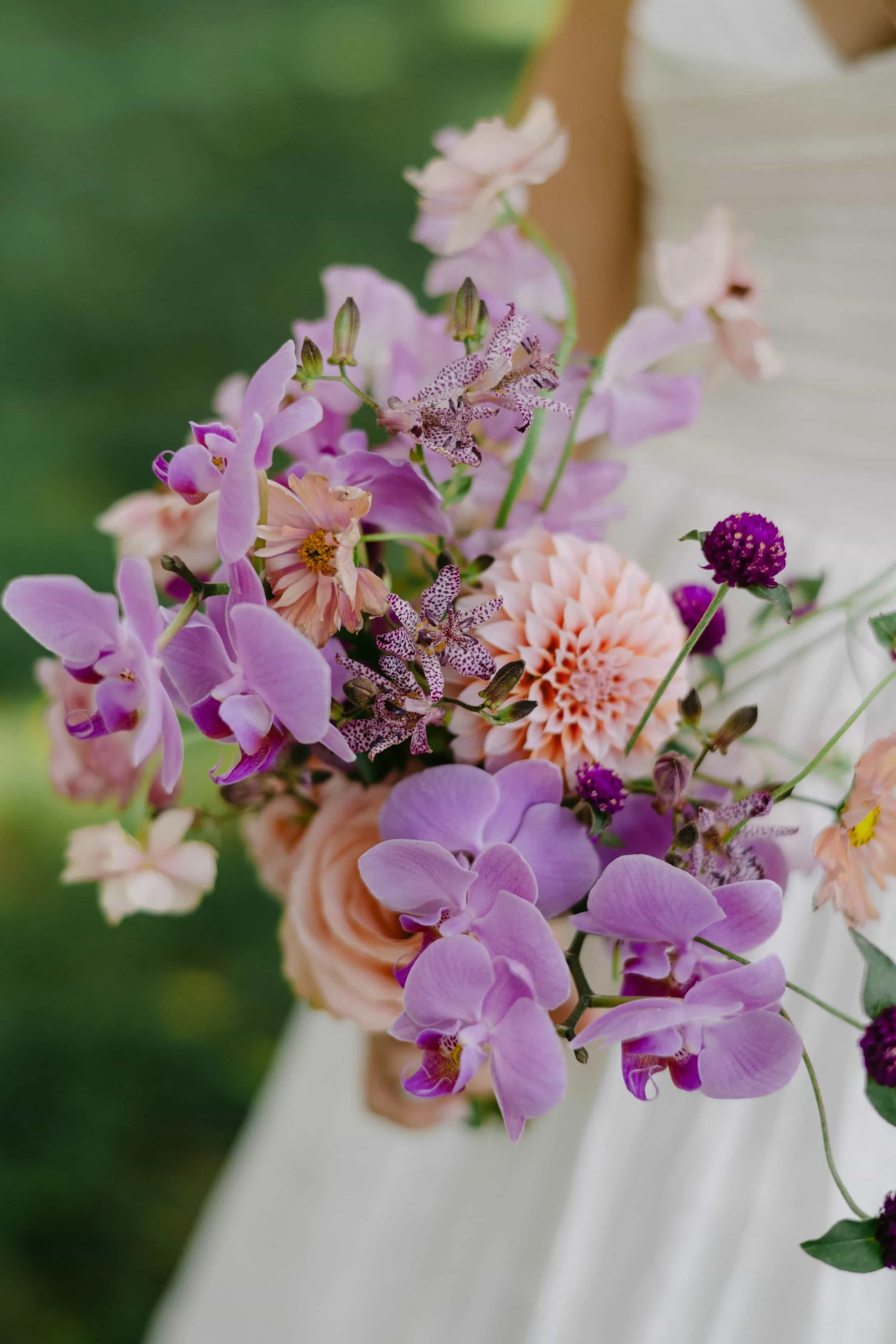 A bouquet of pastel-colored flowers with pink, purple, and peach blossoms, held by someone wearing a white dress.