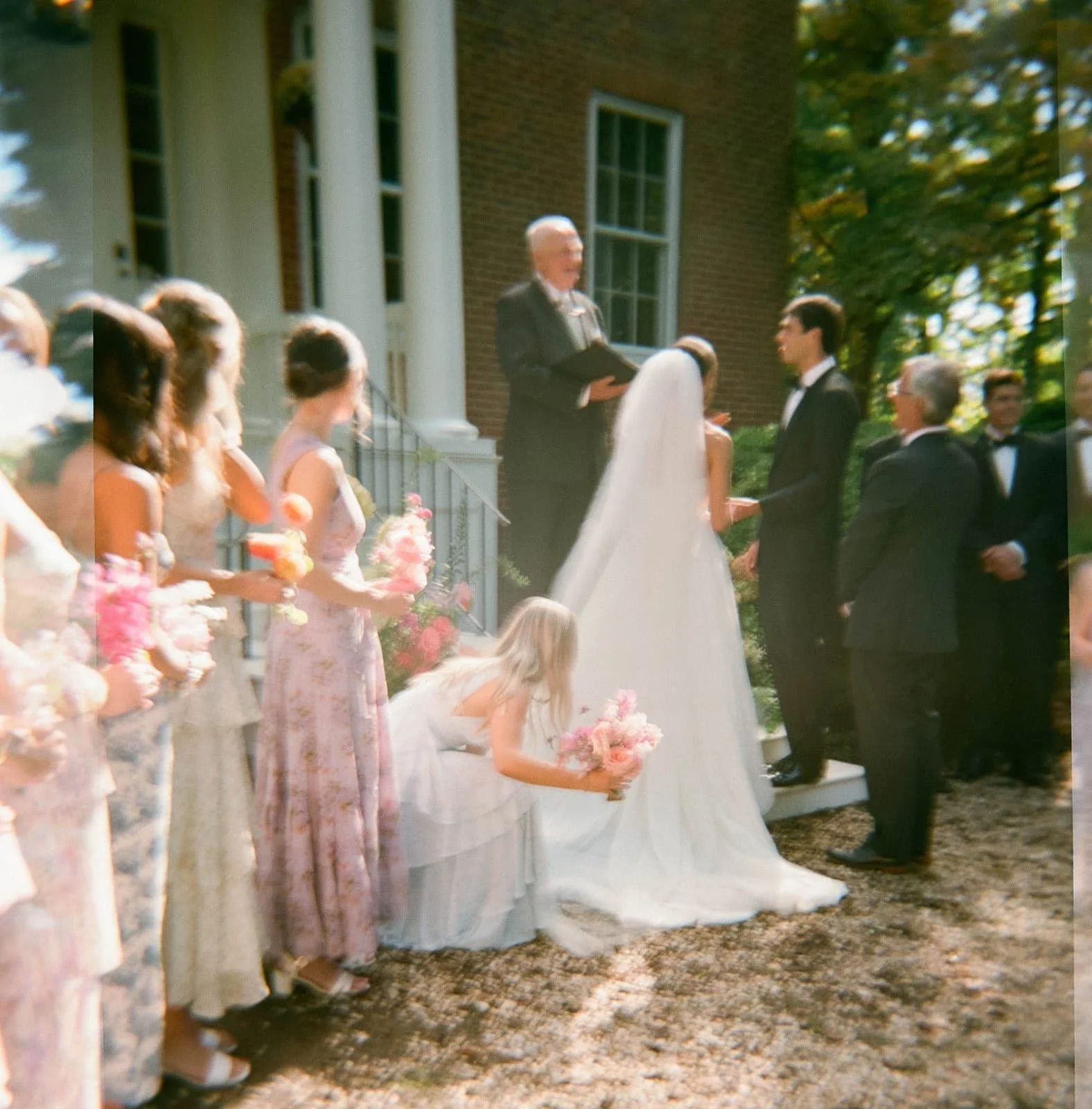A wedding ceremony outdoors with a bride and groom exchanging vows in front of a officiant, surrounded by bridesmaids holding pink bouquets, on a sunny day with green trees and a brick building in the background.