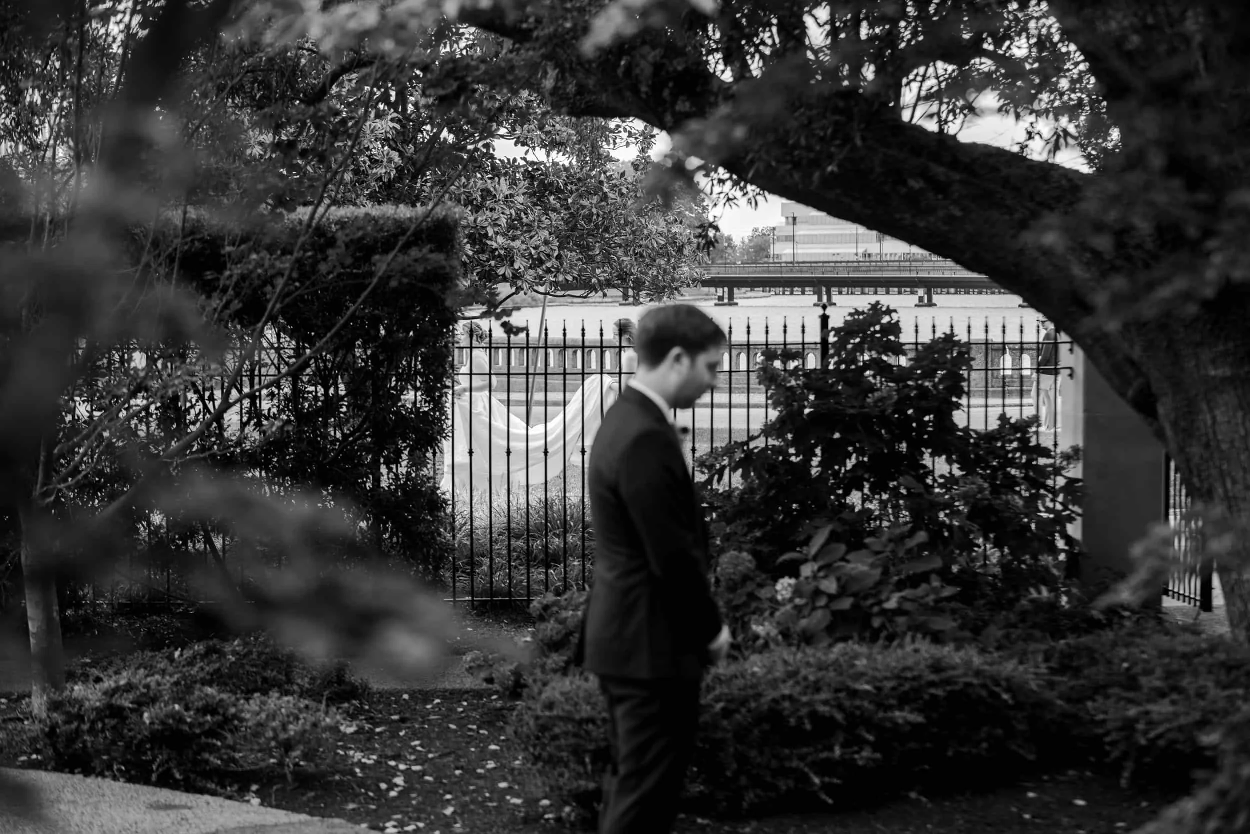 A man in a suit standing in a garden, with trees and bushes around him, and a fence behind him. In the background, there is a large statue and a bridge over a body of water.